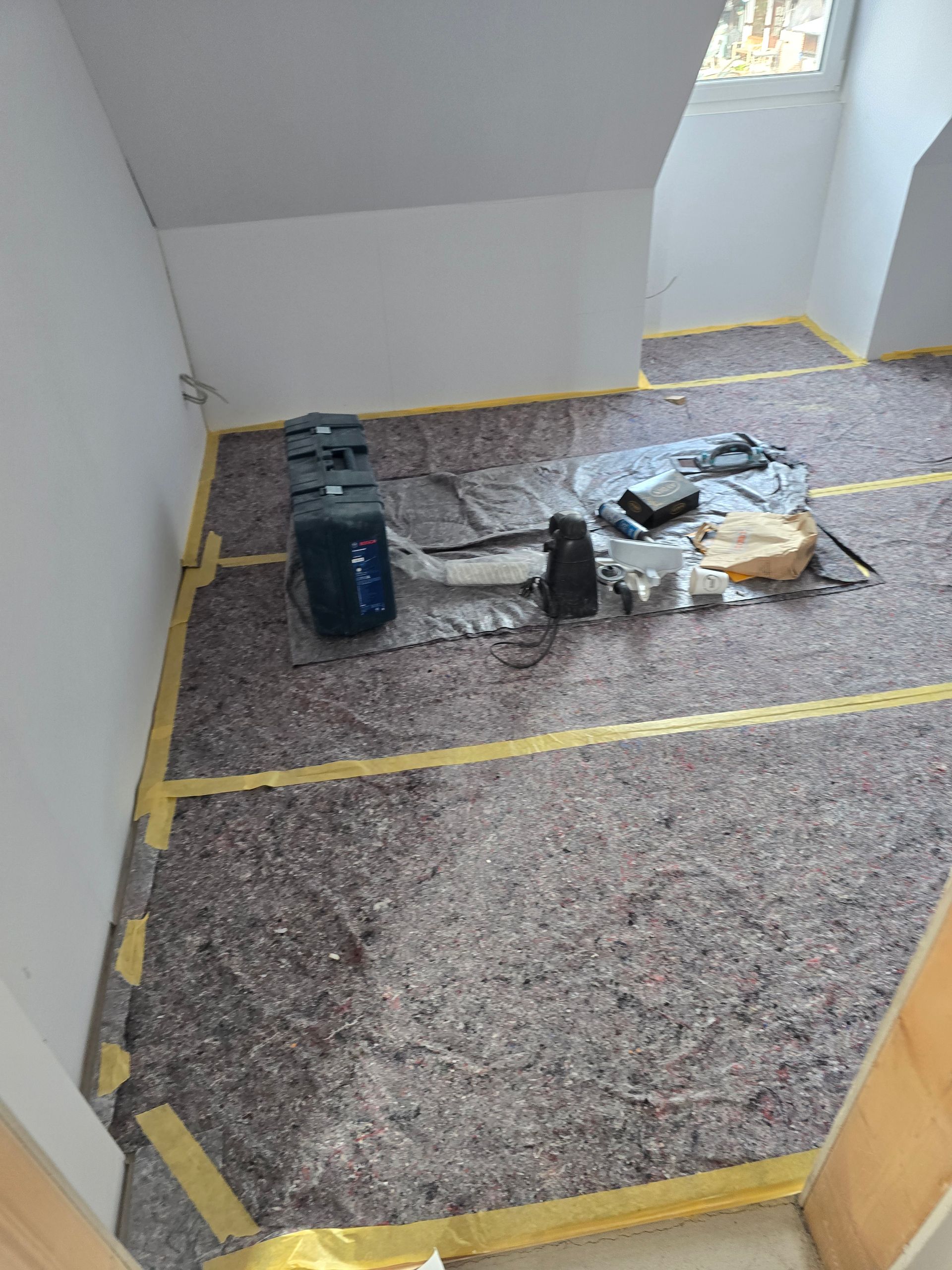 Raum, in dem gerade ein Bodenbelag verlegt wird; graue Dämmung auf dem Boden, Werkzeuge und Material verstreut.