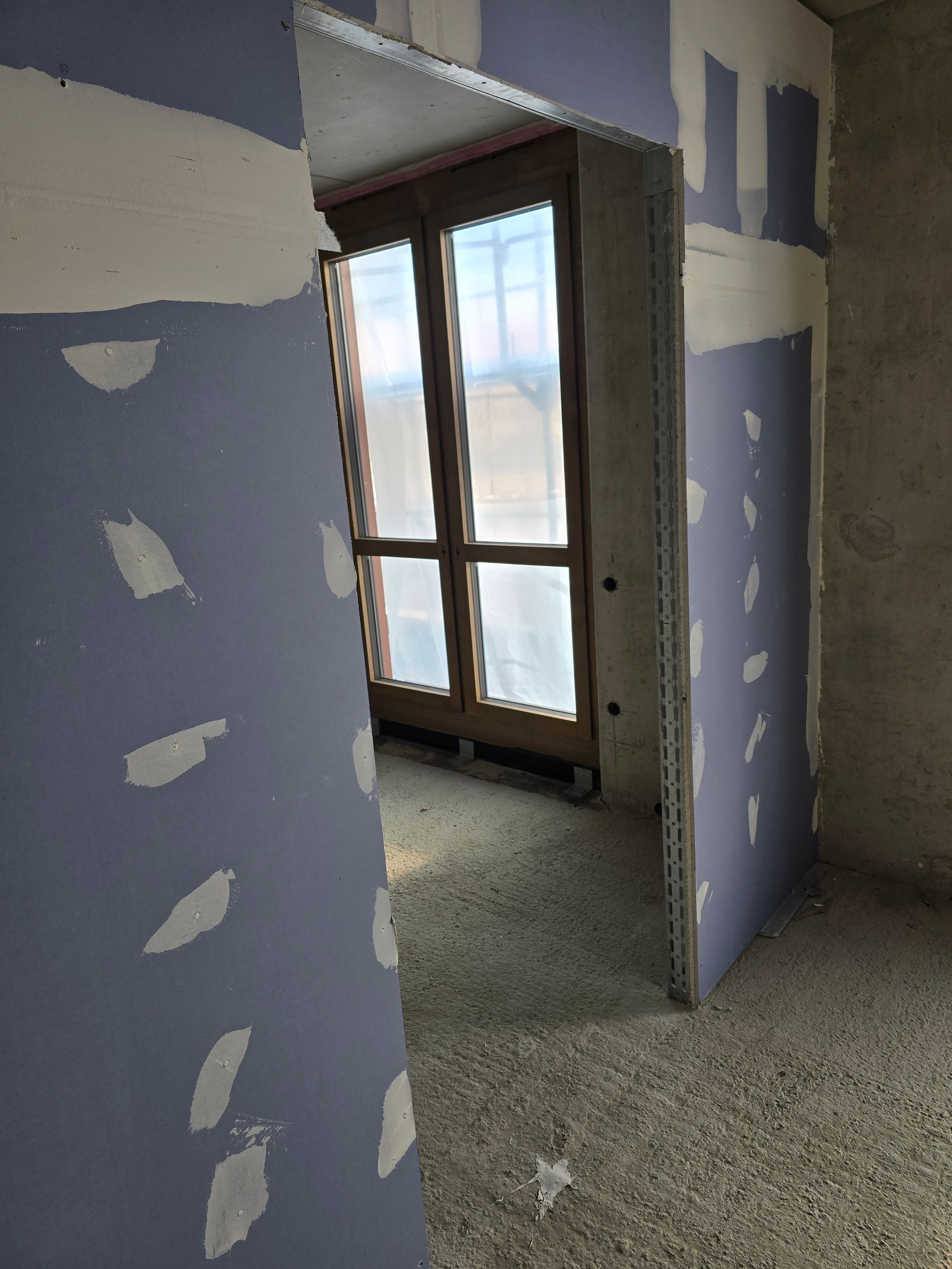 Innenbereich mit unfertigen Trockenbauwänden und Türöffnung, die zu einem Fenster führt.