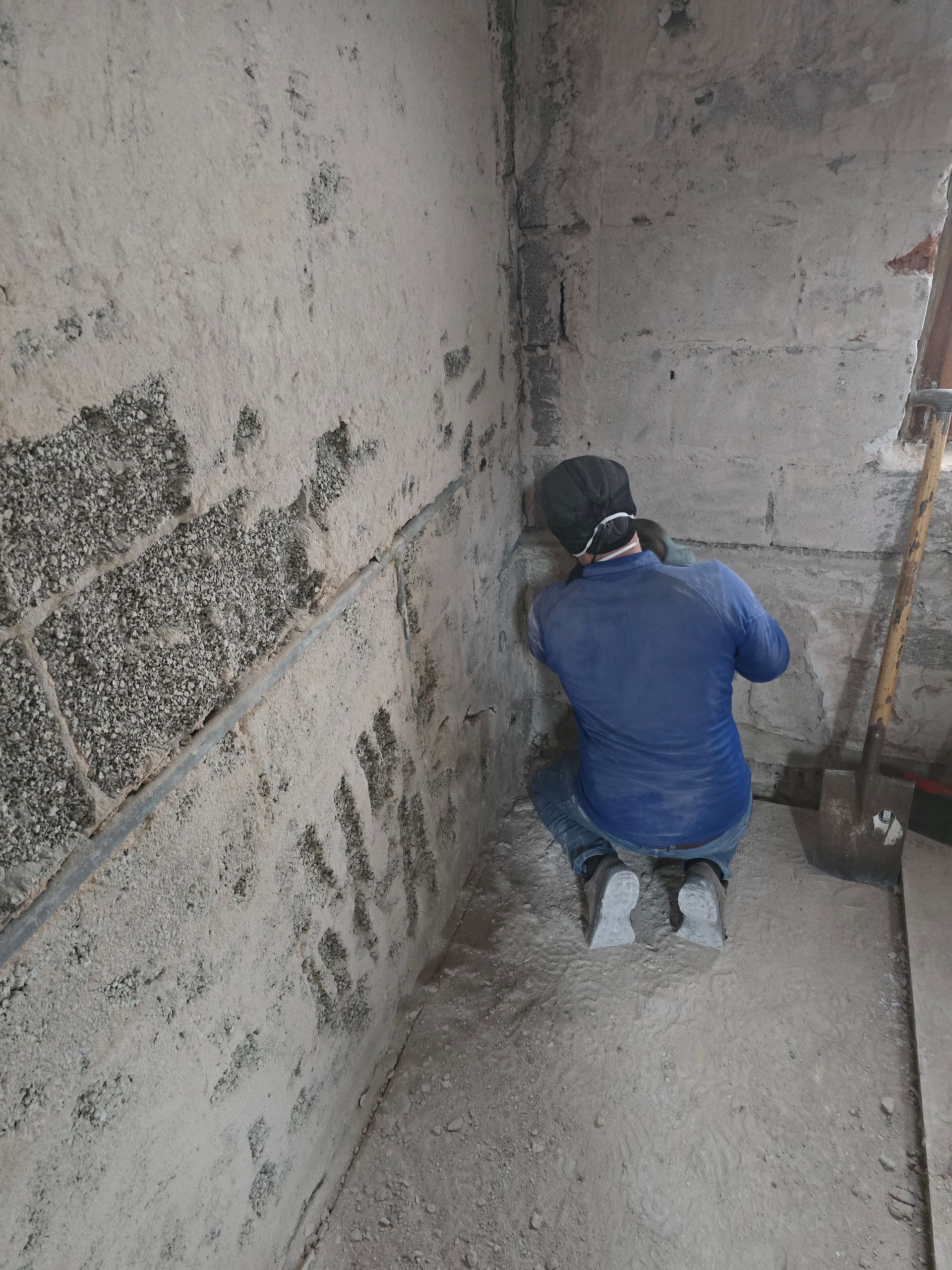 Eine Person kniet und schleift eine Ecke einer Betonwand. Staubige Baustelle, eine Schaufel liegt daneben.