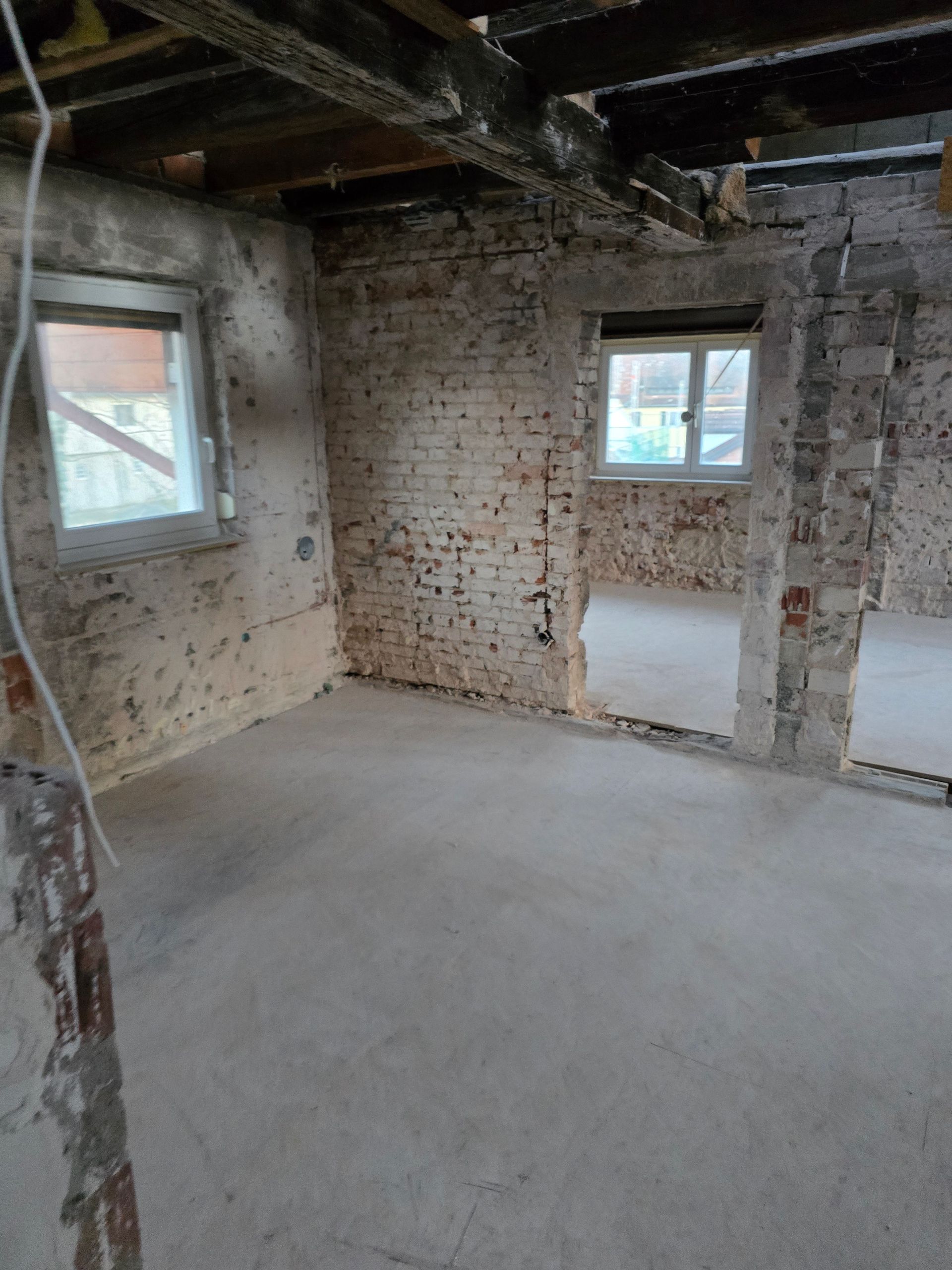 Das Zimmer ist innen baufällig, mit freiliegenden Ziegelwänden, zwei Fenstern, Betonboden und offener Tür zu einem anderen Zimmer.