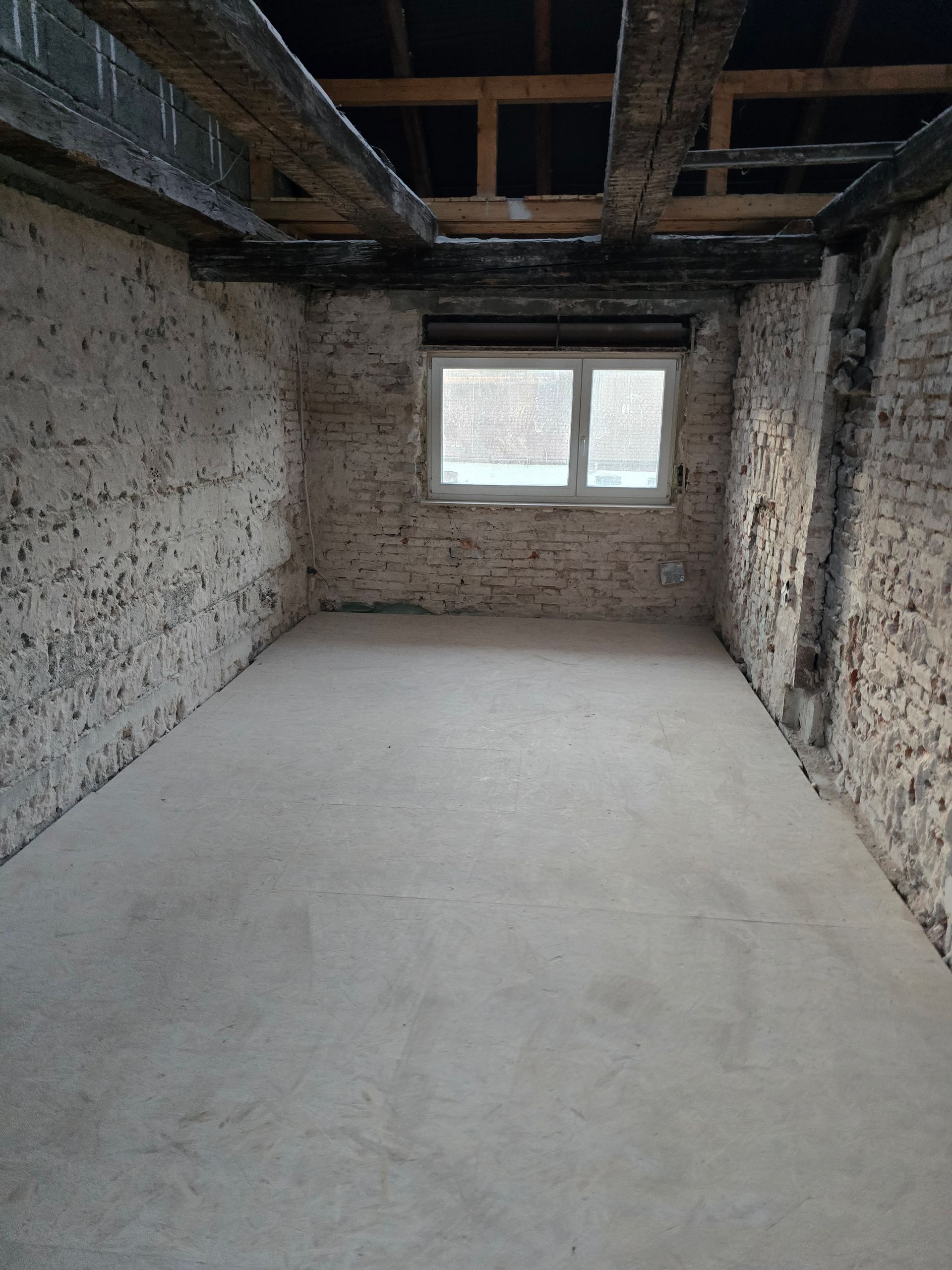 Leerer Raum mit freiliegenden Ziegelwänden, Holzbalken, Betonboden und einem Fenster.