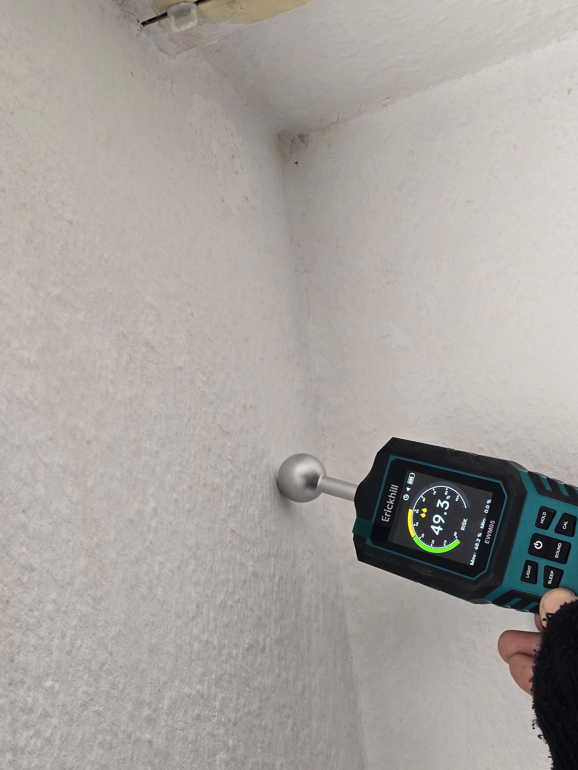 Ein Feuchtigkeitsmessgerät wird an eine weiße Wand in einer Ecke gedrückt und zeigt einen Messwert an.