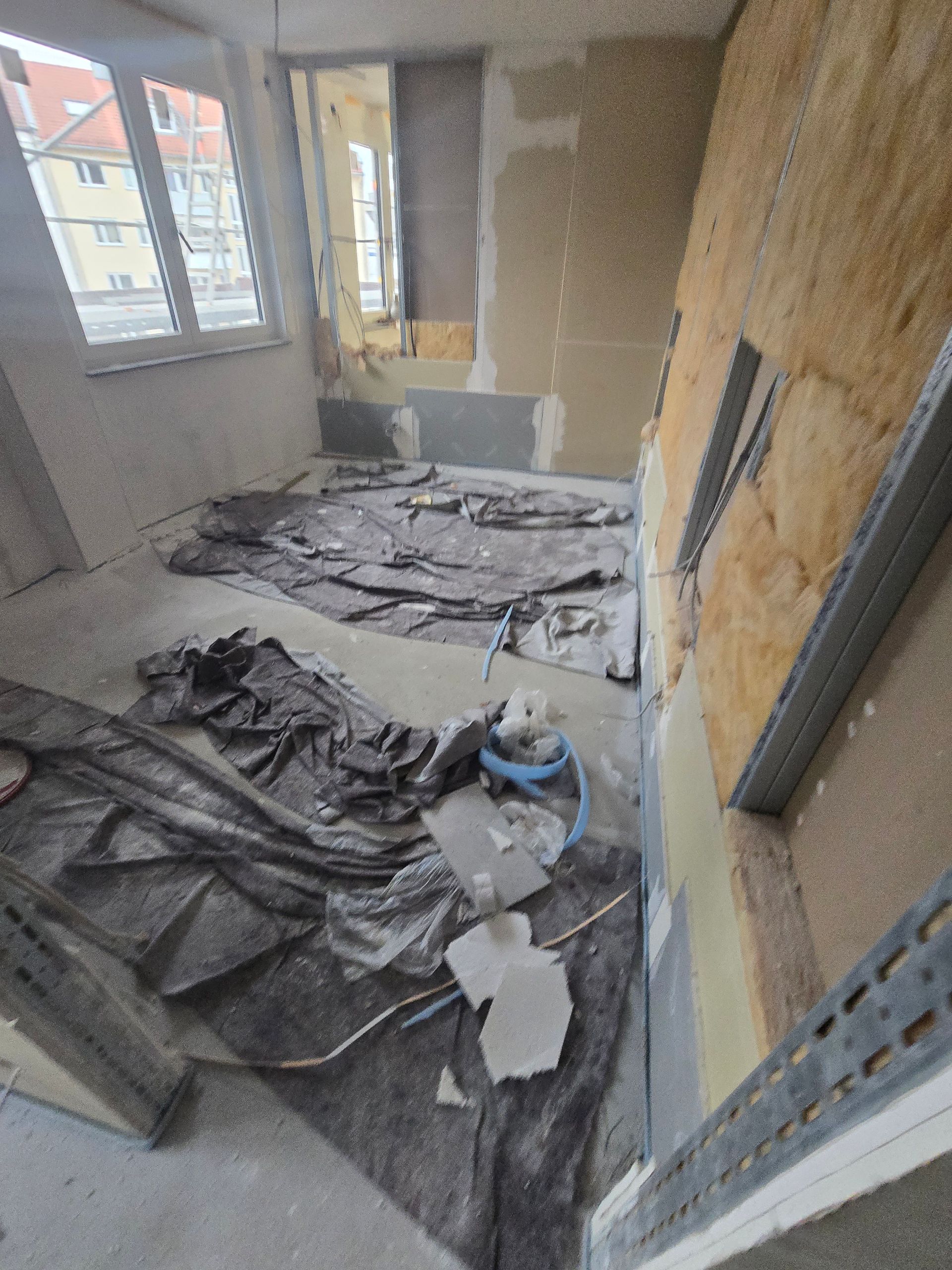 Ein im Bau befindlicher Raum mit freiliegenden Wänden und mit Schutzfolie abgedecktem Boden. Das Fenster ist sichtbar.