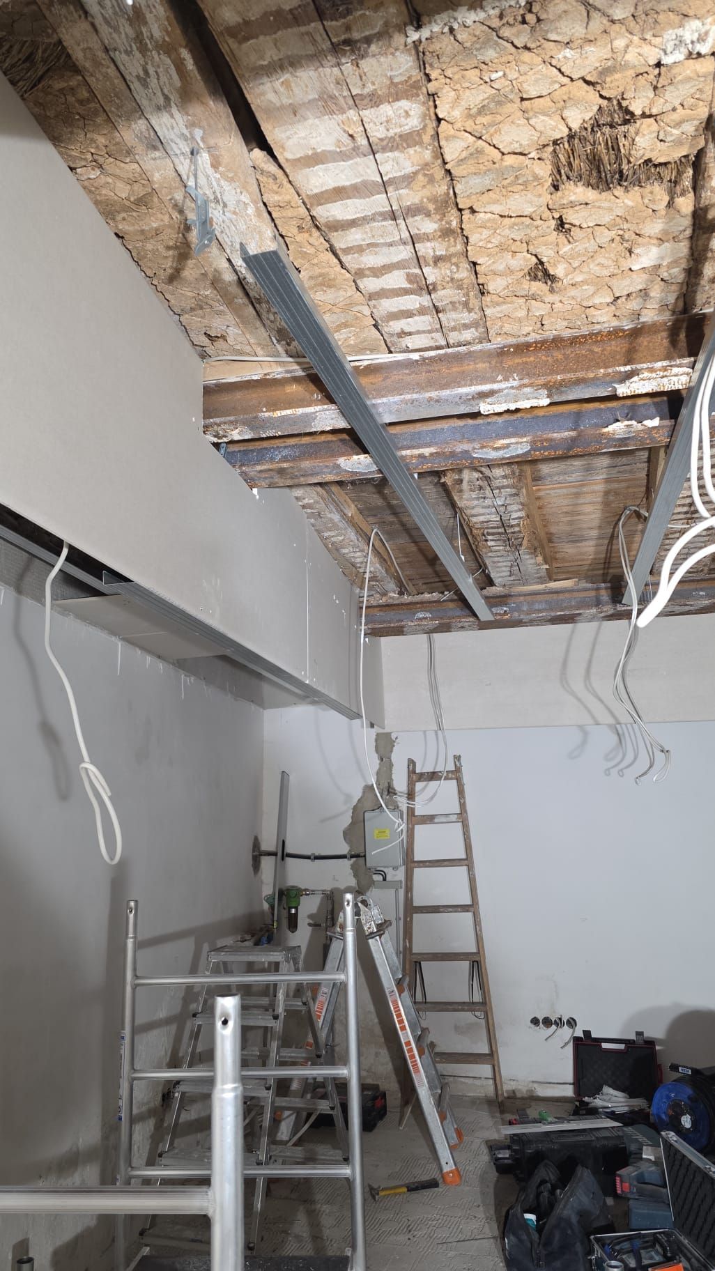 Renovierter Raum: freiliegende Holzbalken an der Decke mit Metallstützen, darunter Baugeräte und eine Leiter.