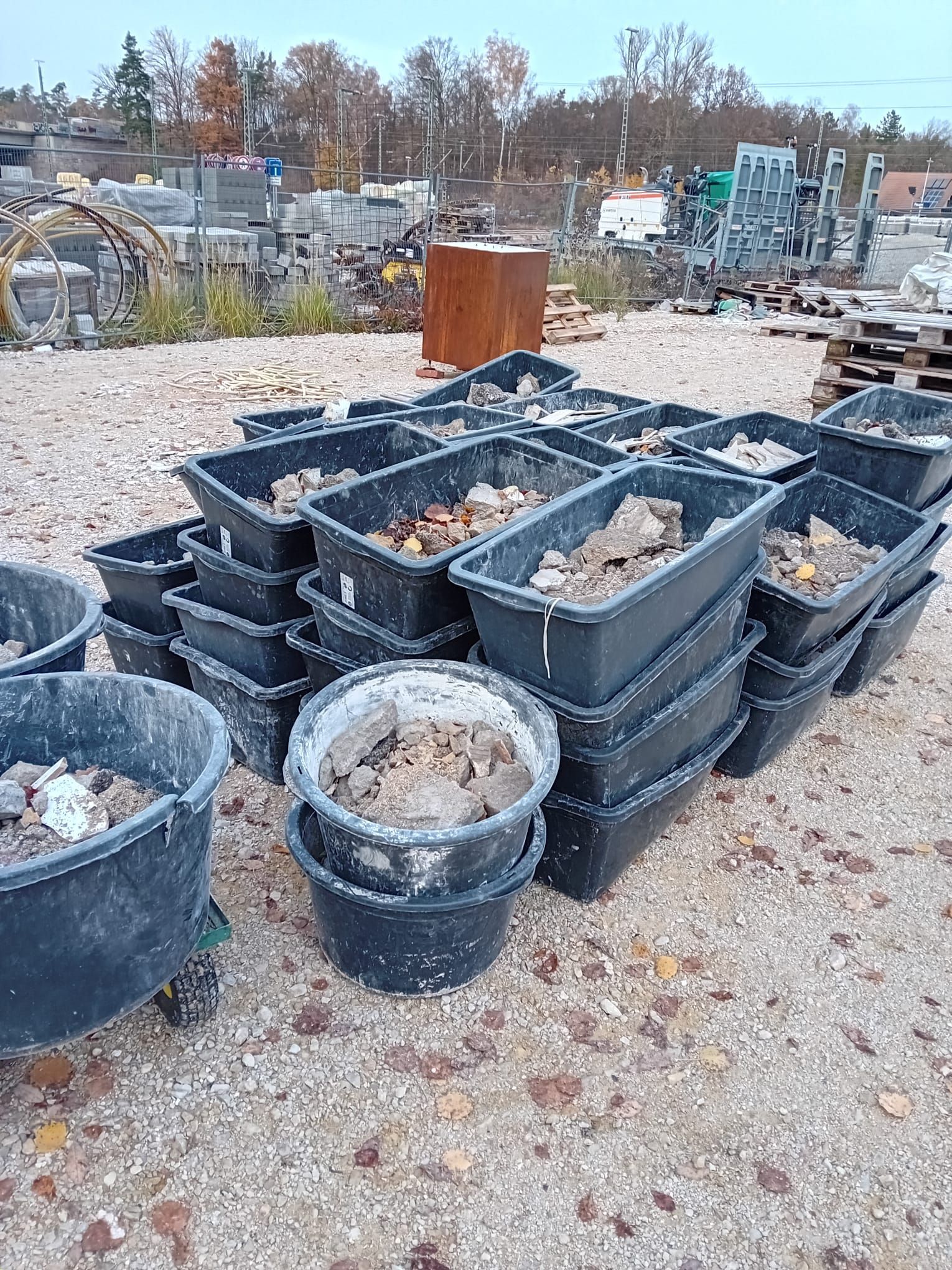 Schwarze Plastikbehälter, teils gestapelt, gefüllt mit Schmutz und Schutt, stehen im Freien auf Kies.
