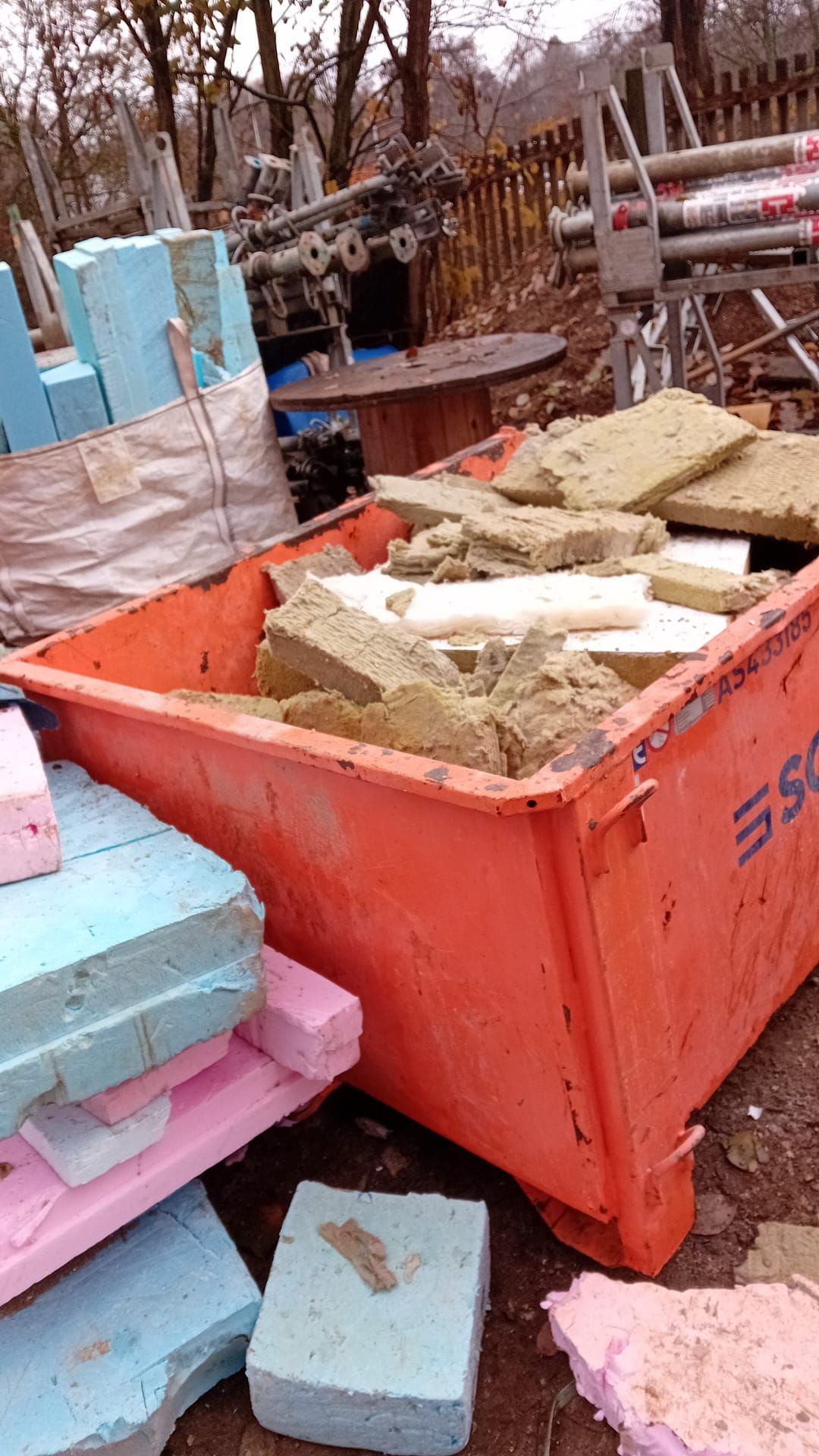 Ein orangefarbener Abfallcontainer, gefüllt mit Dämmmaterial und Schaumstoffblöcken; Baustellenumgebung.