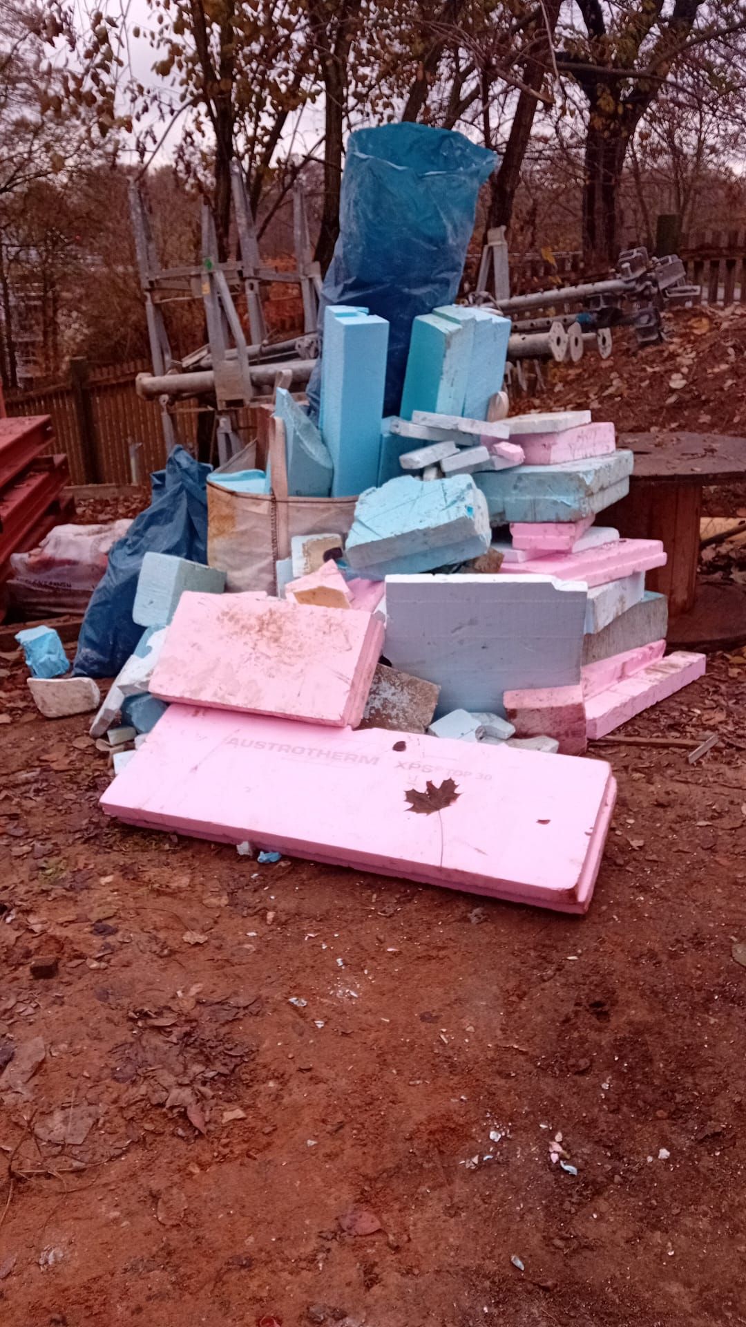 Ein Haufen rosa, blauer und grauer Schaumstoffisolierungsreste liegt auf Erde, im Hintergrund sind Baumaterialien zu sehen.