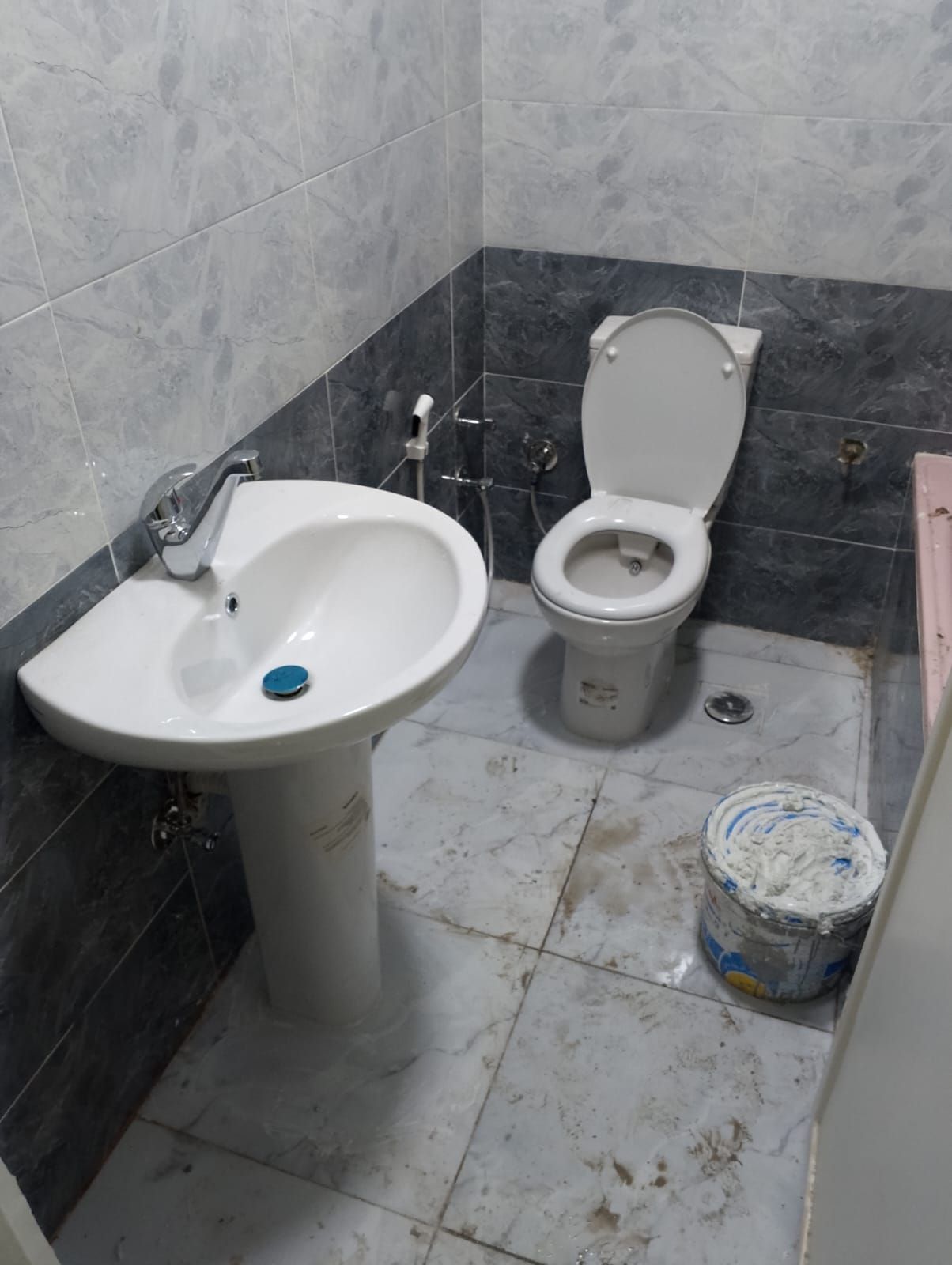 Badezimmer mit weißem Standwaschbecken, Toilette, grauen Fliesen und Farbeimer.