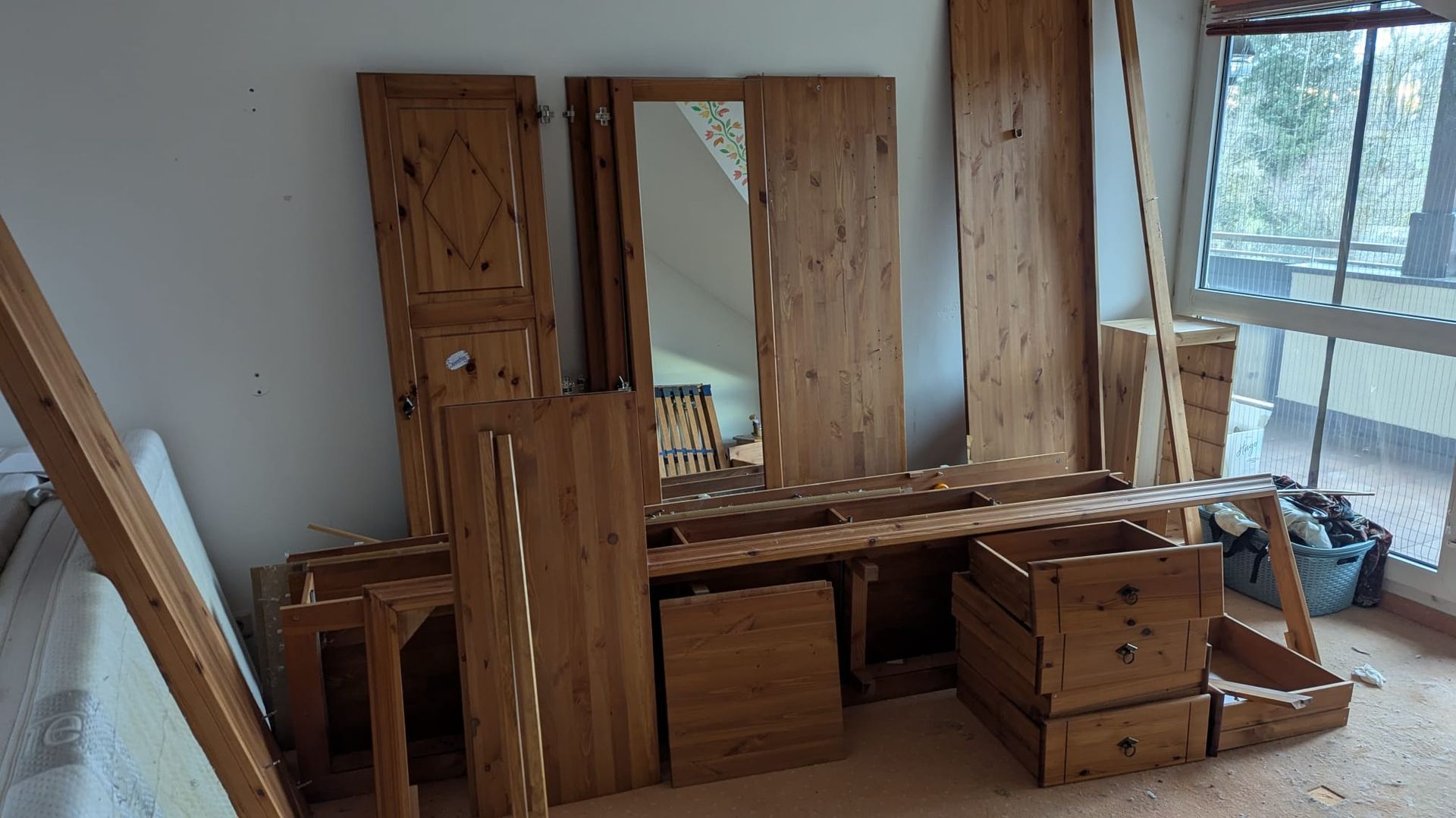 Demontierte Holzmöbelstücke in einem Zimmer mit Fenster, darunter ein Schminktisch, Schranktüren und Schubladen.