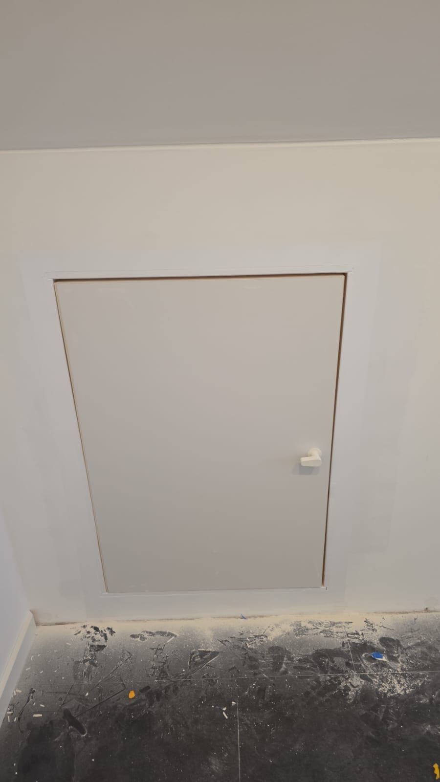 Weiße, rechteckige Zugangsklappe in einer weißen Wand mit einem kleinen weißen Türknauf, vermutlich ein Serviceeingang.