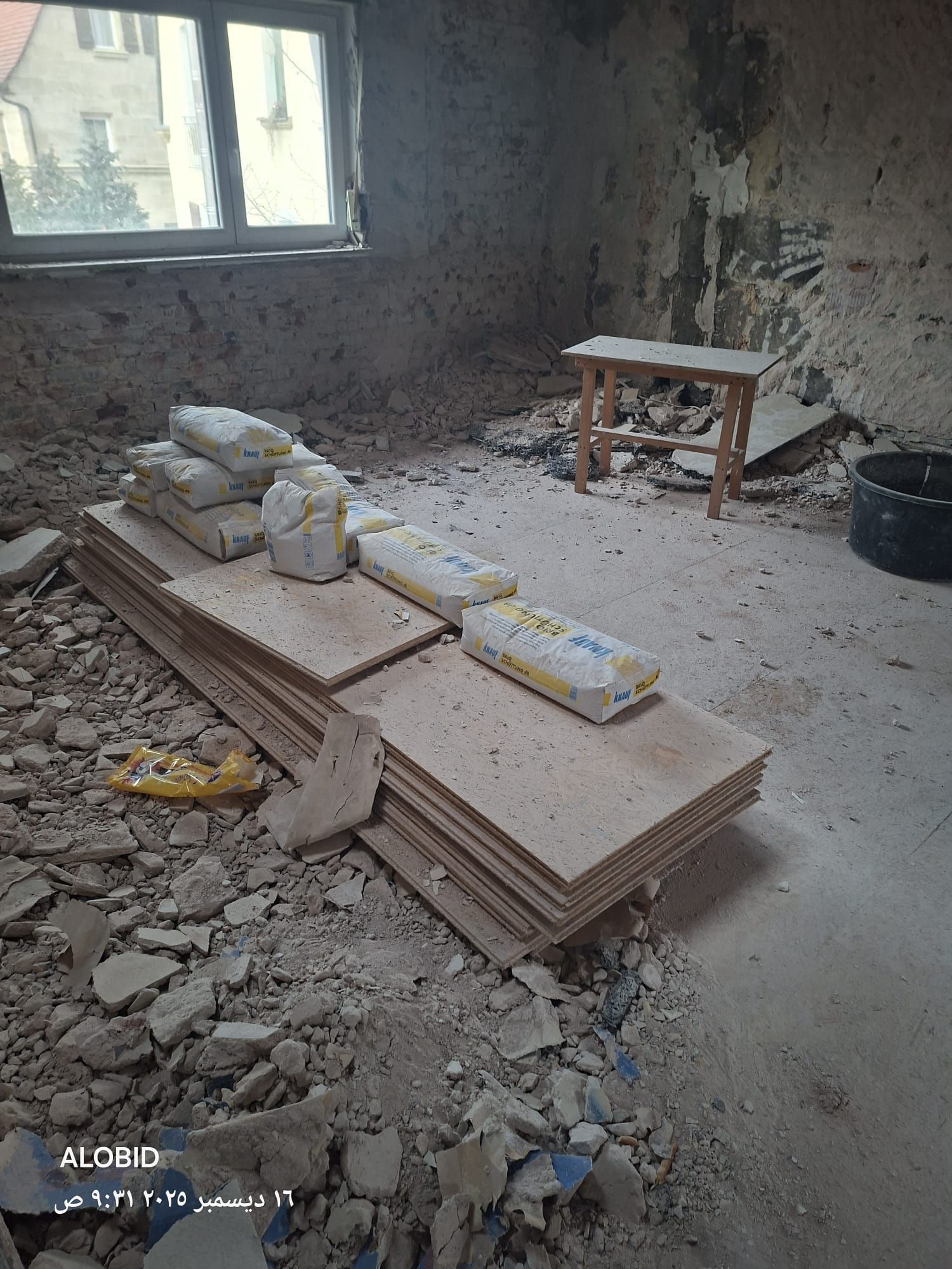 Ein Raum, der gerade renoviert wird, mit gestapelten Fliesen, Säcken mit Baumaterial, einem kleinen Hocker und Bauschutt.