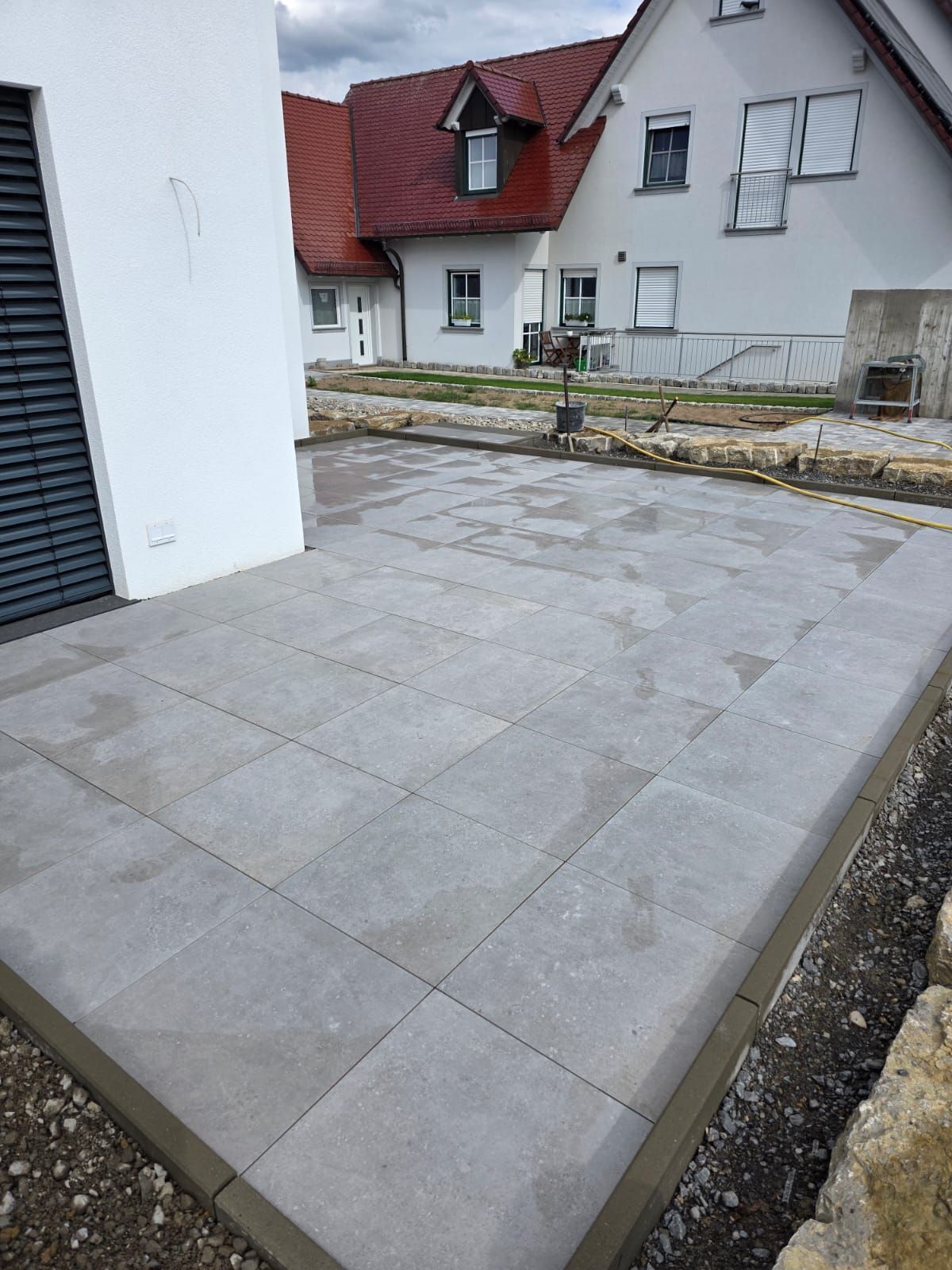 Neu verlegte graue Terrassenfliesen, eingefasst von dunkelgrauem Beton, neben einem weißen Haus.
