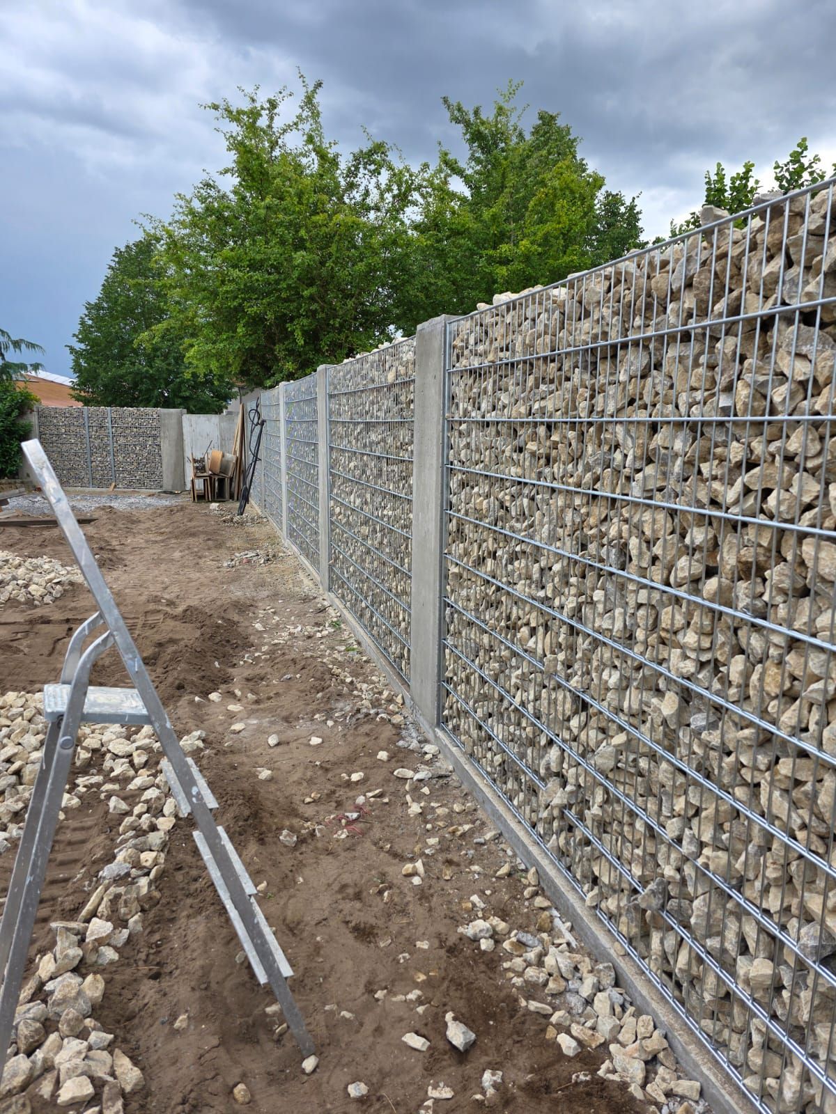 Gabionenzaun im Bau; Drahtgeflecht gefüllt mit Steinen, Betonpfosten und umgebender Erde.