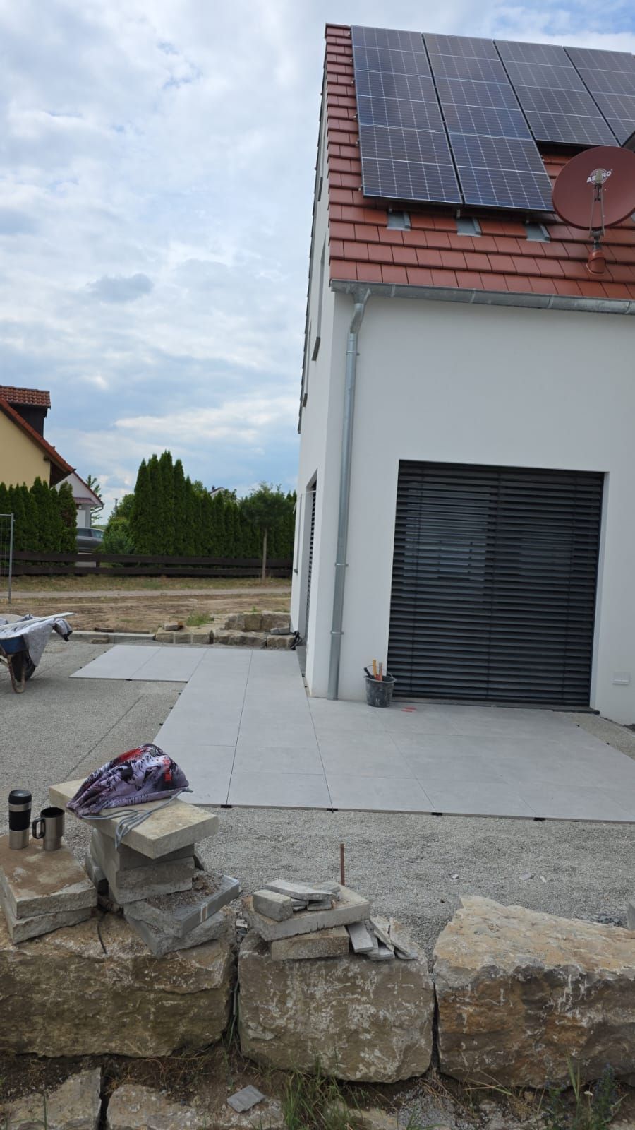 Außenansicht eines Hauses mit Garage, Solaranlage und Steingarten. Graue Betonterrasse, bewölkter Himmel.