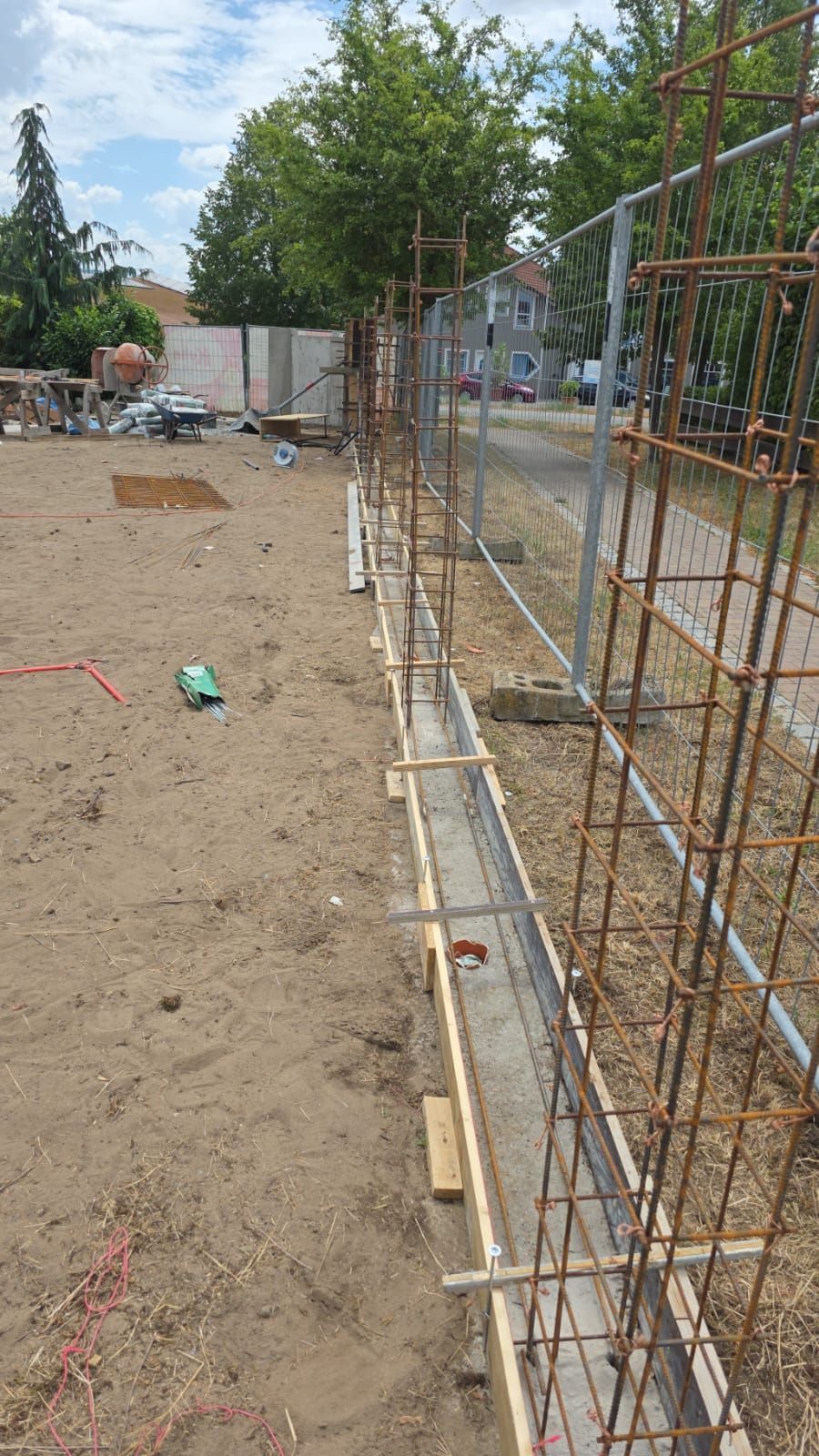 Baustelle mit Stahlbetongerüst neben einem Betonfundament, einem Zaun und sandigem Untergrund.