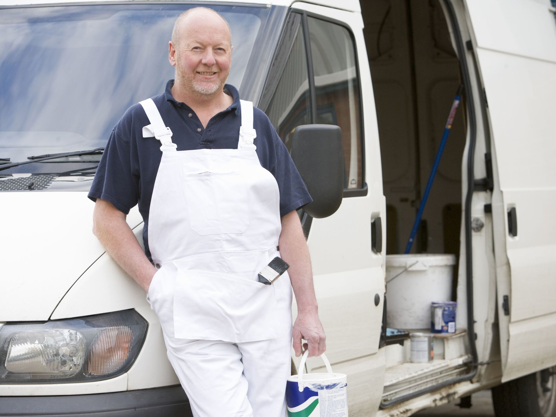 Ein Maler in weißem Overall lehnt lächelnd an einem weißen Lieferwagen und hält einen Farbeimer in der Hand.