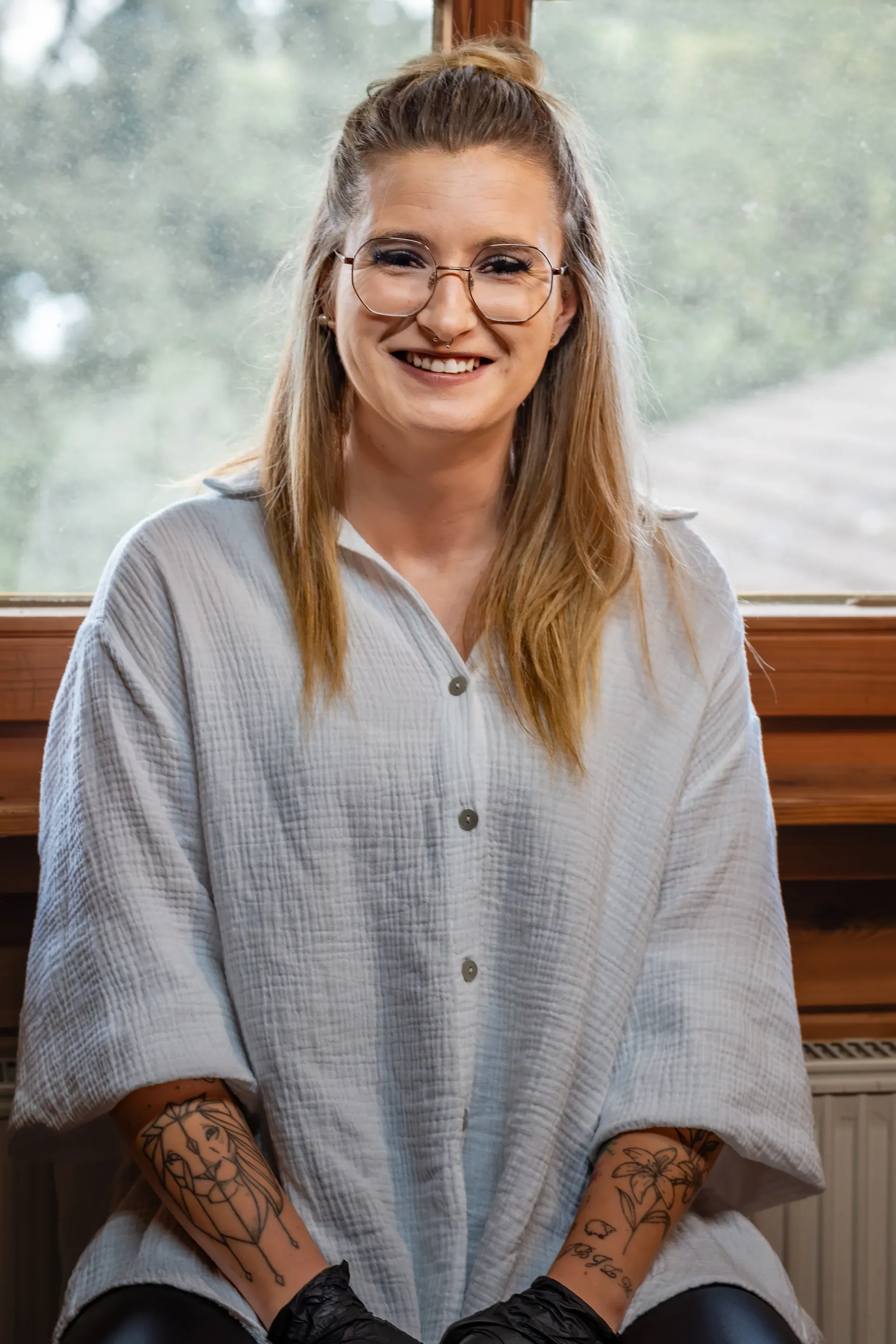 Eine lächelnde Person in einem hellen, strukturierten Oberteil mit Tätowierungen an beiden Unterarmen sitzt vor einem Fenster.