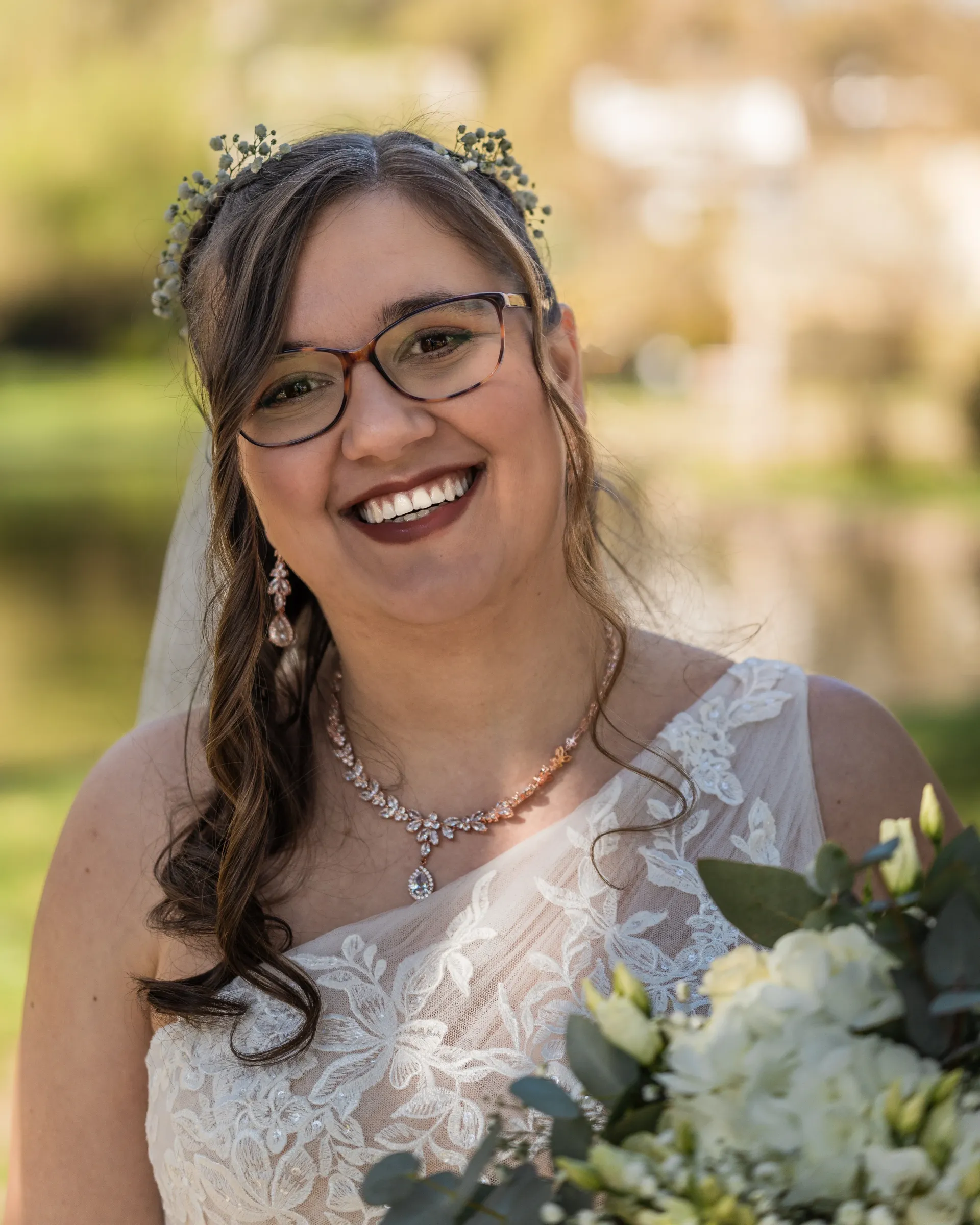 Eine lächelnde Person in einem weißen Brautkleid aus Spitze und mit Brille, die einen Blumenhaarschmuck und eine Halskette trägt und einen Blumenstrauß hält.