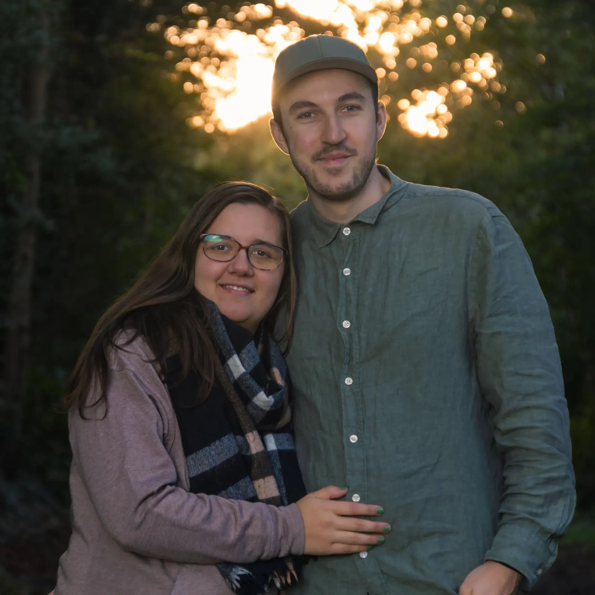 Nelli und Jonathan stehen im Wald und blicken frontal in die Kamera. Im Hintergrund ist ein Sonnenuntergang zu sehen.
