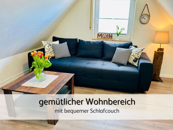 Wohnzimmer der FeWo Lütte Krabbe in Bensersiel mit Blick auf das gemütliche Schlafsofa. Im Vordergrund ist ein Couchtisch mit frischen Blumen