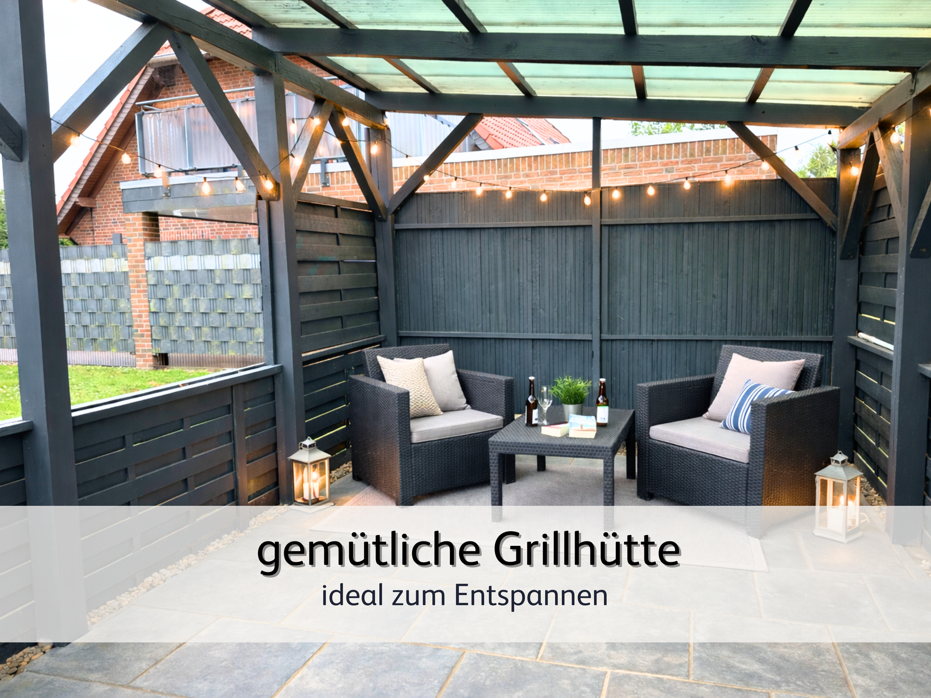 die gemütliche Gartenhütte der Ferienwohnung Lütte Krabbe. schwarze Rattan-Sessel mit gemütlichem Polster sind links und rechts zu sehen. Neben den Sesseln steht eine warm leuchtende Laterne. In der Mitte steht ein Tisch mit einer Blume. An der Wand leuchtet eine Lichterkette warm