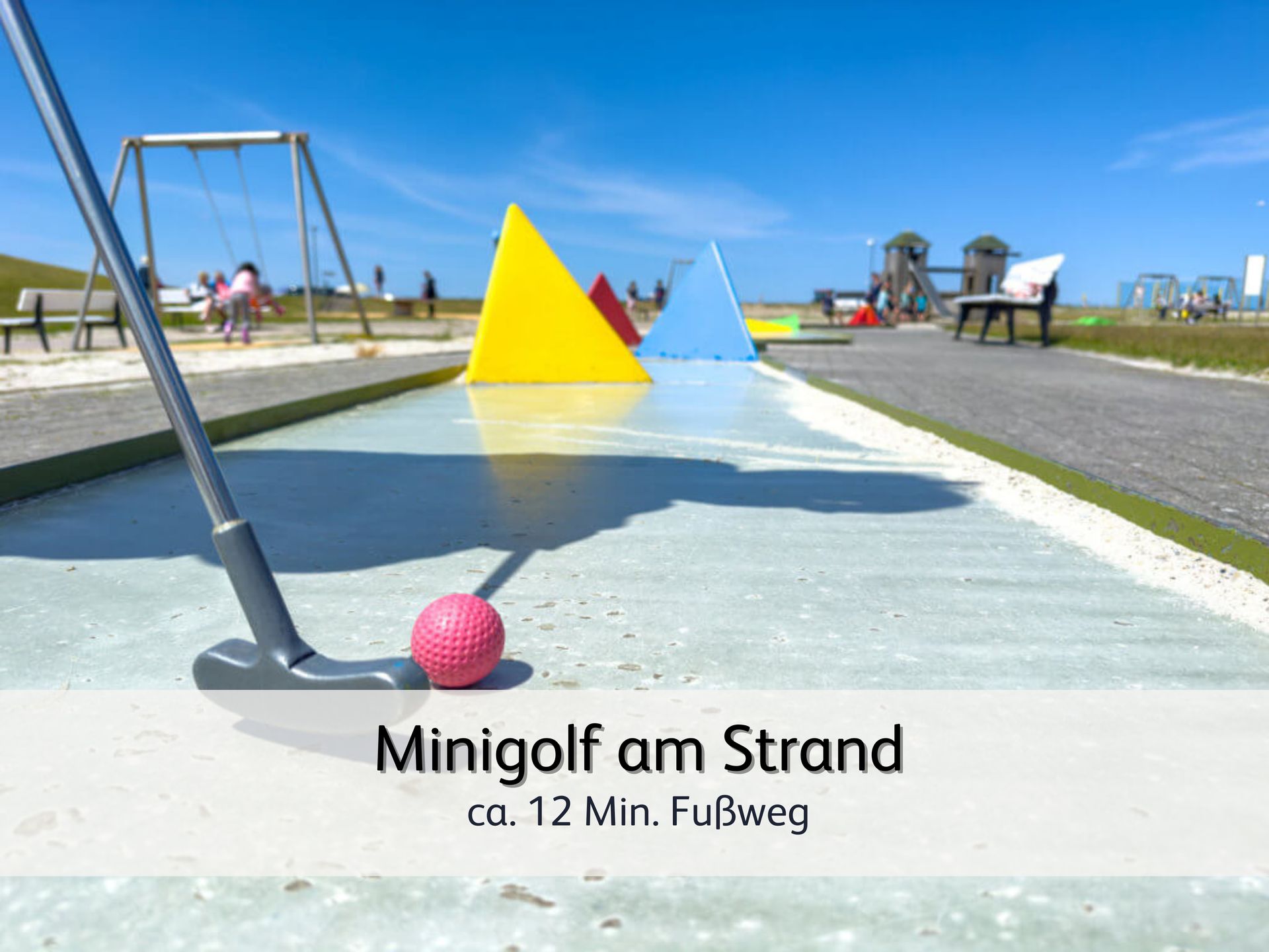 Eine gepflegte Minigolfanlage direkt am Strand von Bensersiel. Im Vordergrund ist ein roter Golfball vor einem Golfschläger. Im Hintergrund sind vor dem blauen Himmel 2 bunte Hindernisse zu sehen