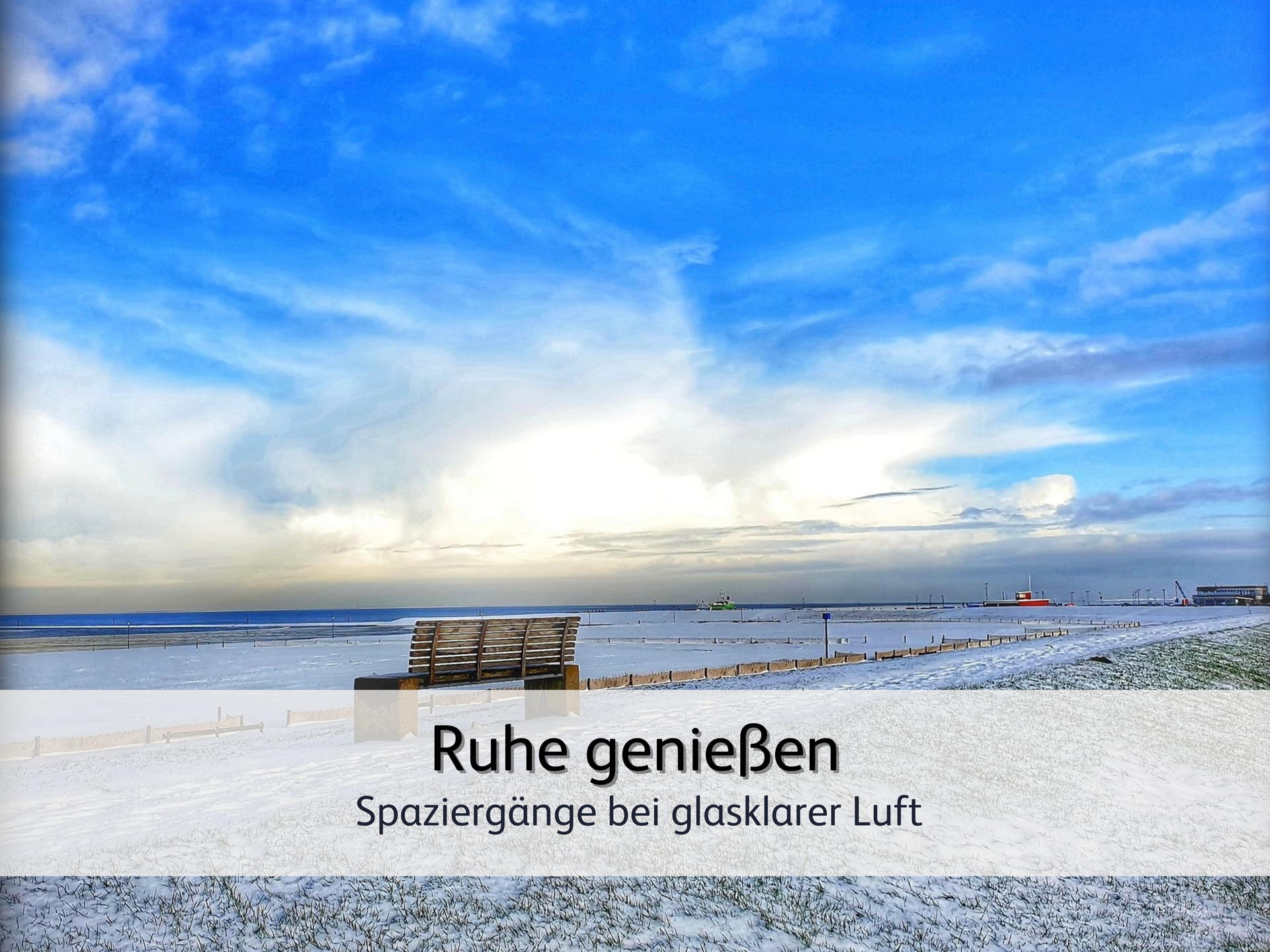 Blick auf den winterlichen Strand von Bensersiel. Im Vordergrund befindet sich eine verschneite Bank auf dem Deich. Von Dort schaut man über den schneebedeckten Strand auf das Meer