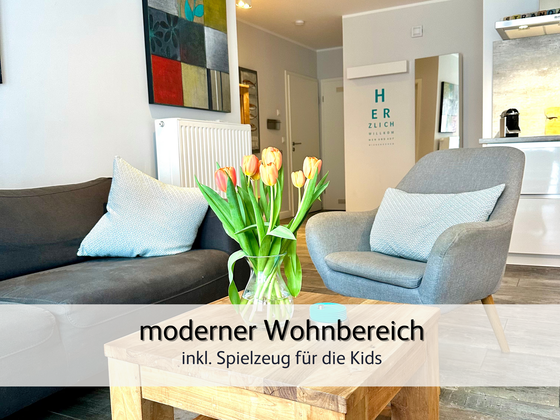 Wohnzimmer der FeWo StrandART in Norddeich mit Blick auf das gemütliche Sofa und den Sessel. Im Vordergrund sind frische Blumen