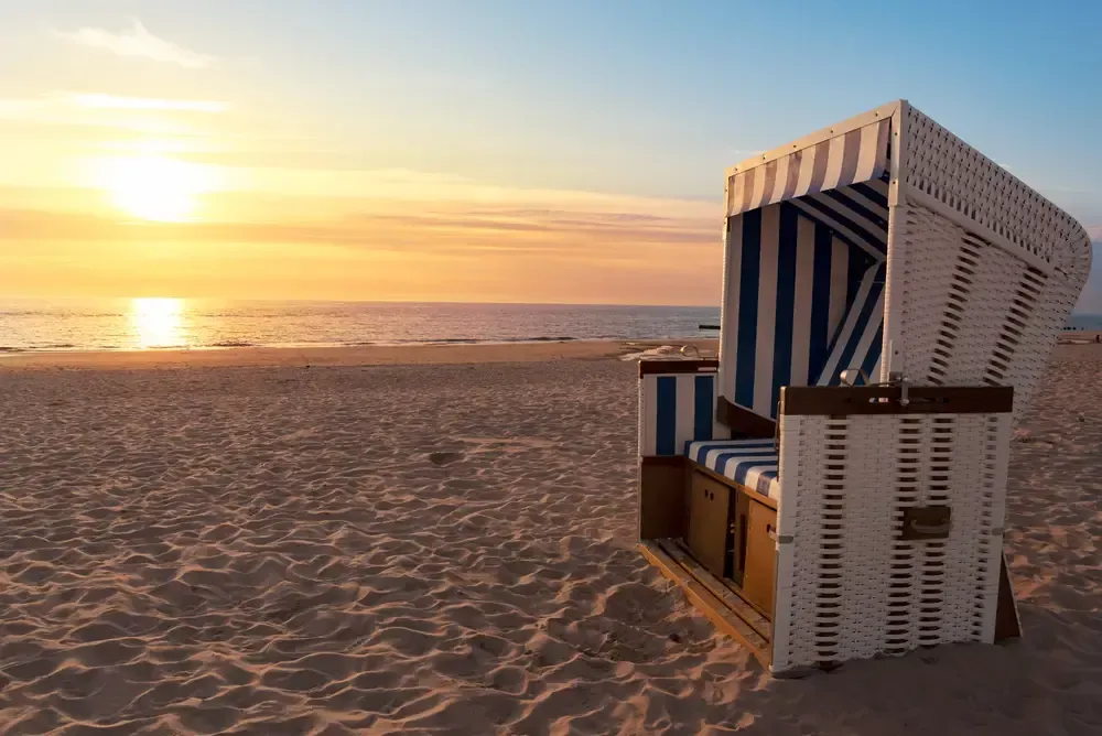 der sommerliche Sandstrand. Im Vordergrund ist ein blau-weißer Strandkorb zu sehen, wie sie an der deutschen Nordseeküste bekannt sind. Im Hintergrund geht die Sonne langsam hinter der Nordsee unter