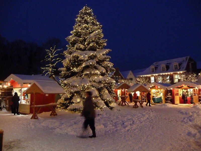 der verschneite Weihnachtsmarkt bei Abenddämmerung. Im Vordergrund steht ein verschneiter, beleuchteter Tannenbaum. im Hintergrund sind die hell und festlich beleuchteten Hütten zu sehen