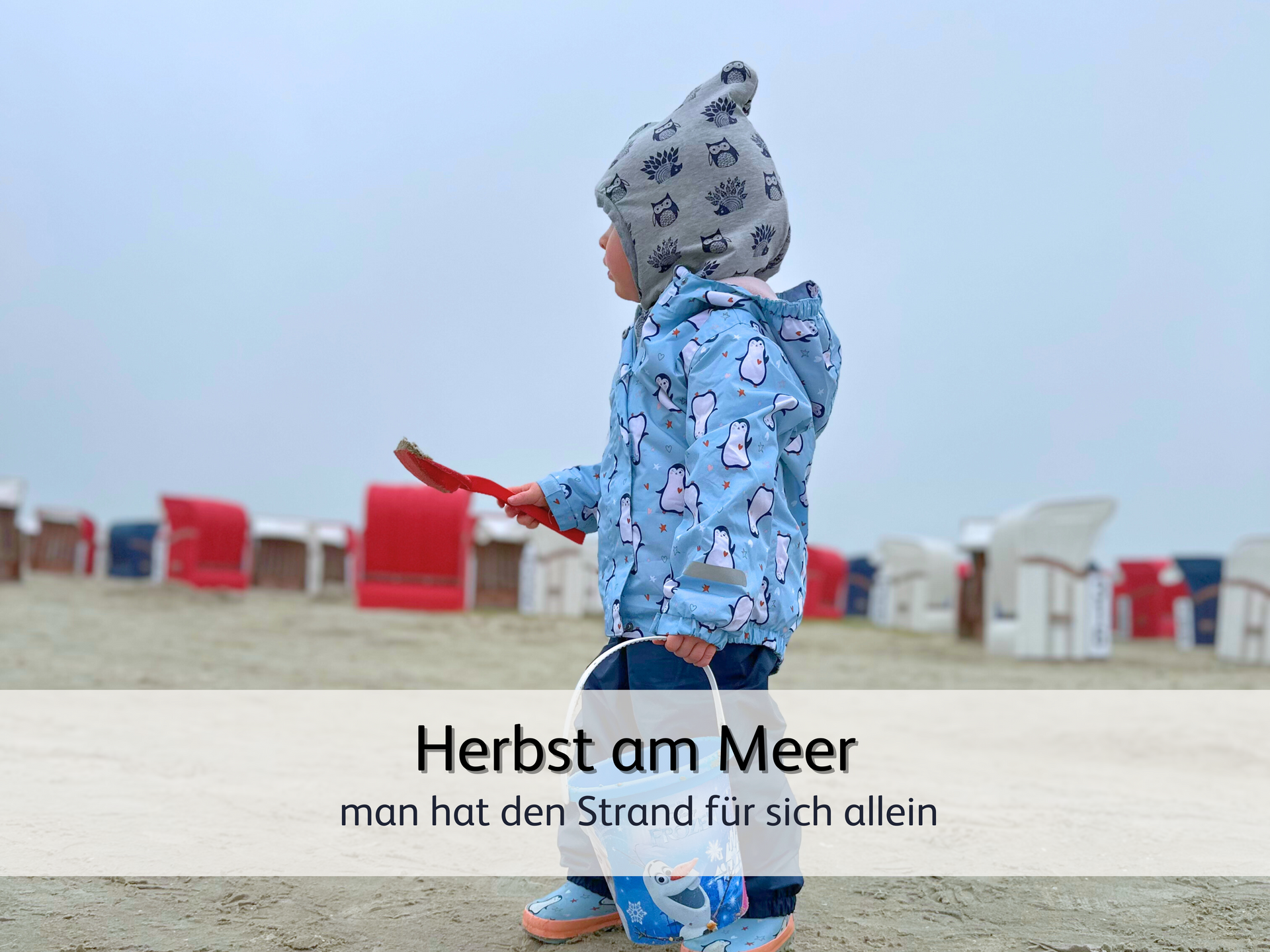 Ein Kind, das in Regenjacke am menschenleeren Strand spielt. In der Hand hält es eine Schaufel und einen Eimer