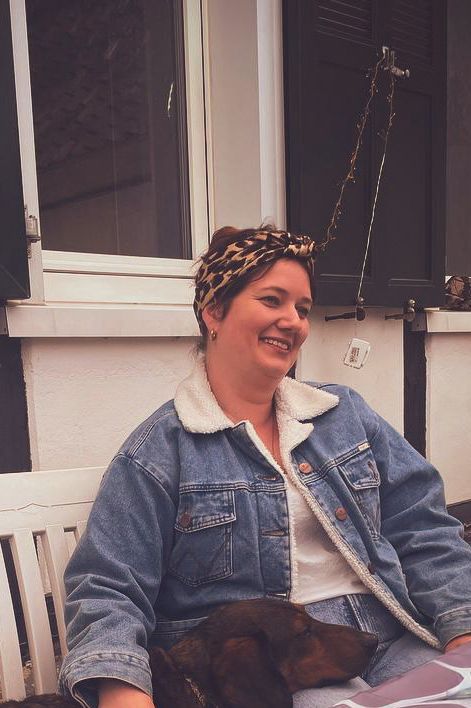 Frau in Jeansjacke und Leopardenmuster-Stirnband lächelt, während sie draußen sitzt, Hund auf dem Schoß.