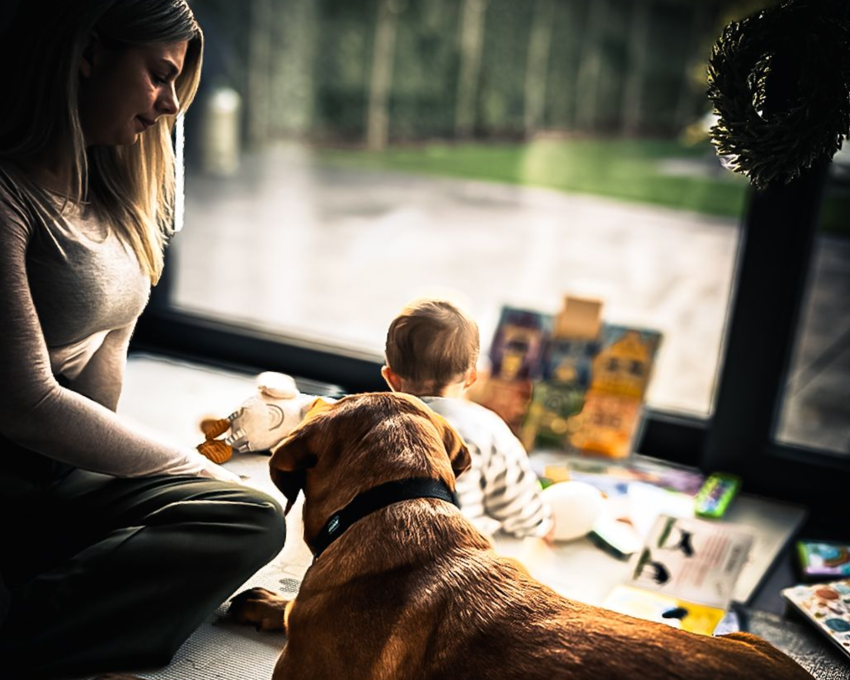 Frau und Baby betrachten Bücher auf dem Boden am Fenster; Hund liegt daneben.