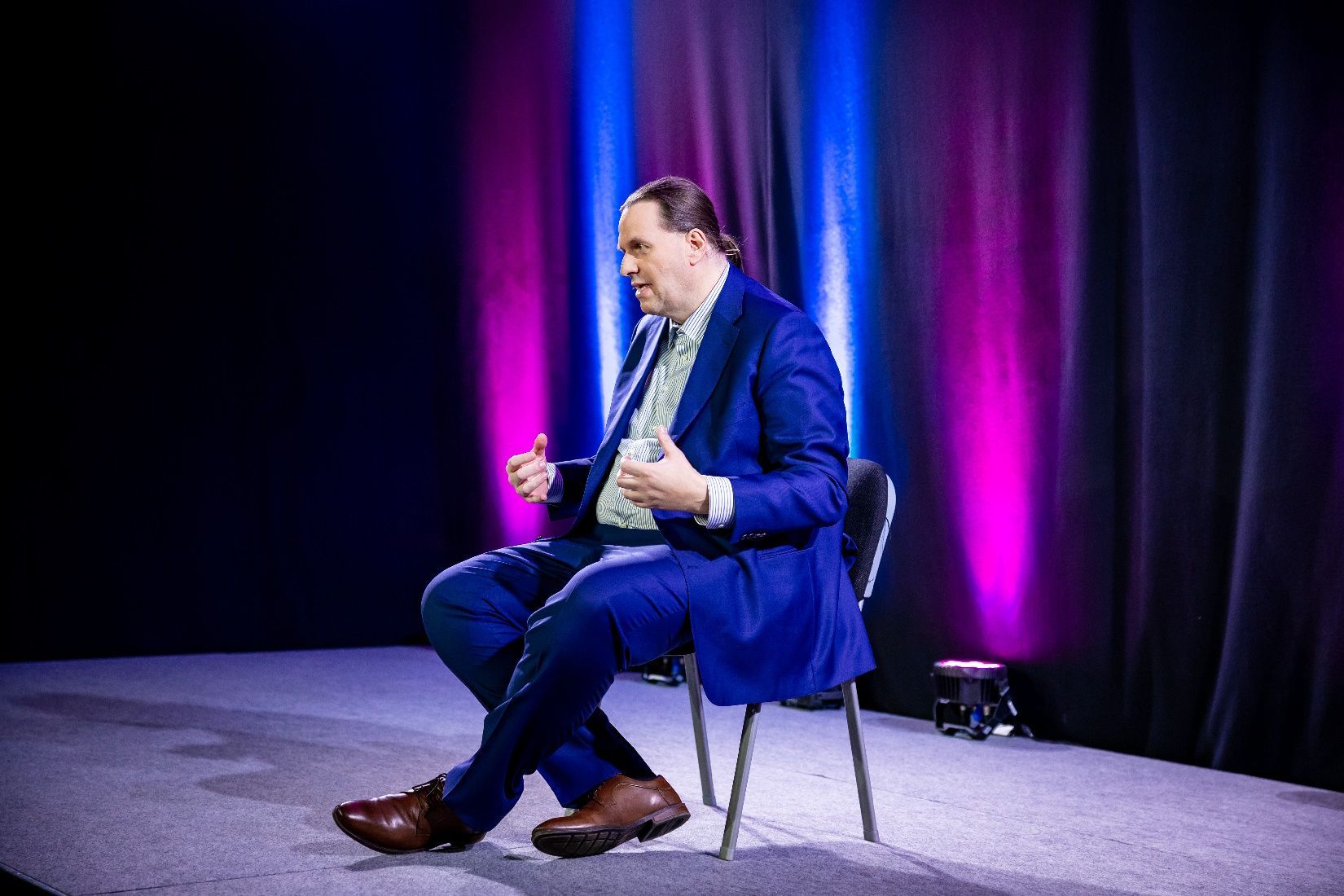 Ein Mann im blauen Anzug sitzt auf der Bühne, gestikuliert mit den Händen und spricht.