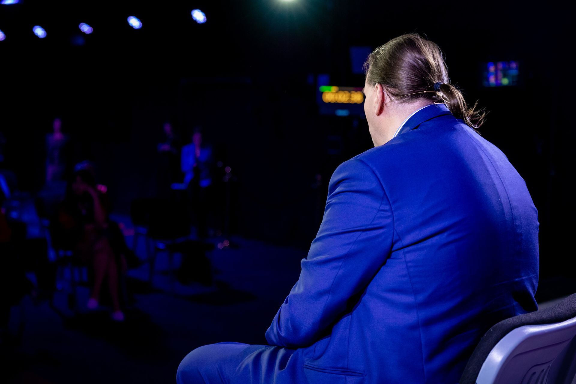 Mann im blauen Anzug, mit Pferdeschwanz, sitzt auf der Bühne und blickt in einem dunklen Raum zum Publikum.
