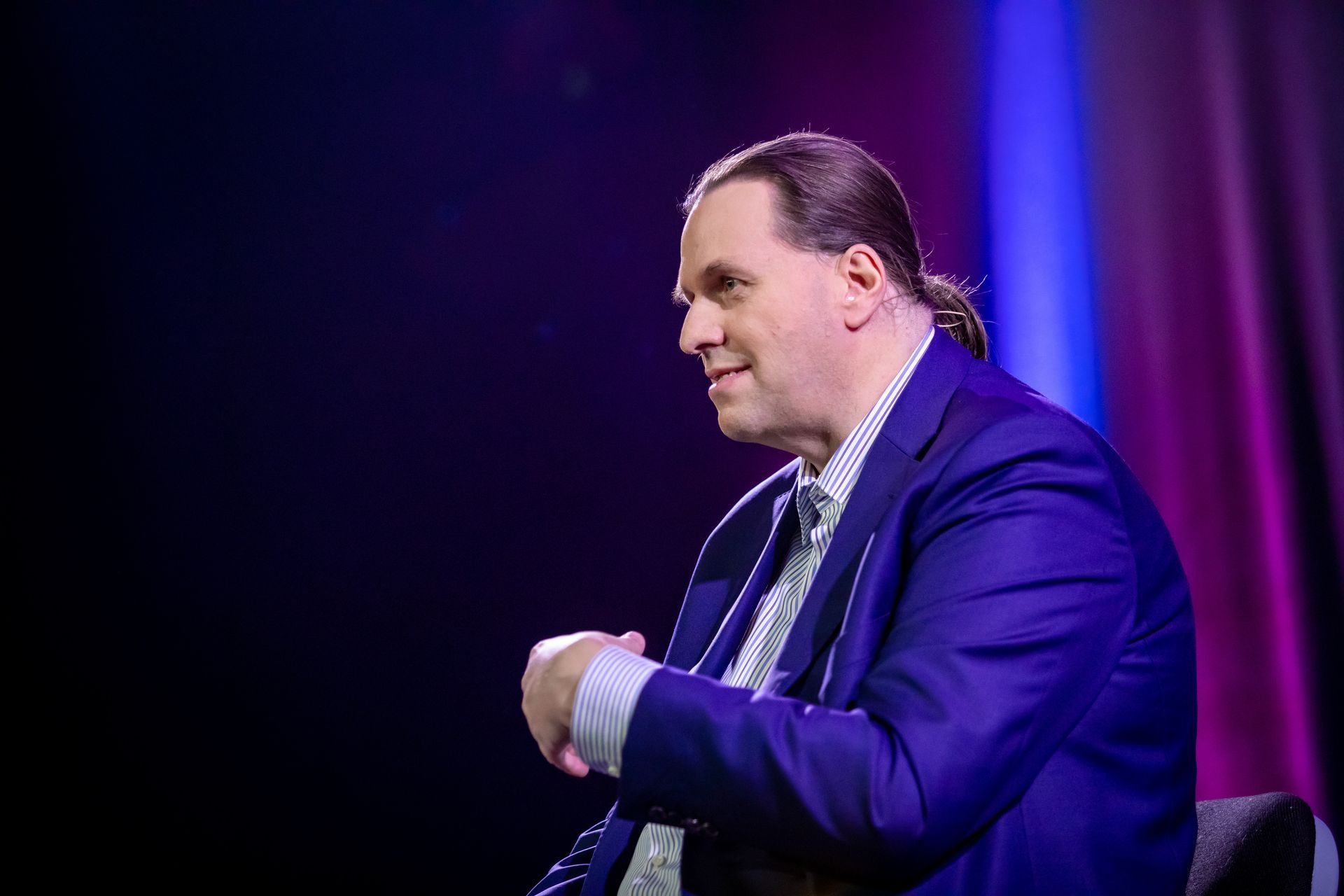 Ein Mann in blauem Blazer und mit Pferdeschwanz lächelt, während er vor dunklem Hintergrund und violetter Beleuchtung auf einer Bühne sitzt.