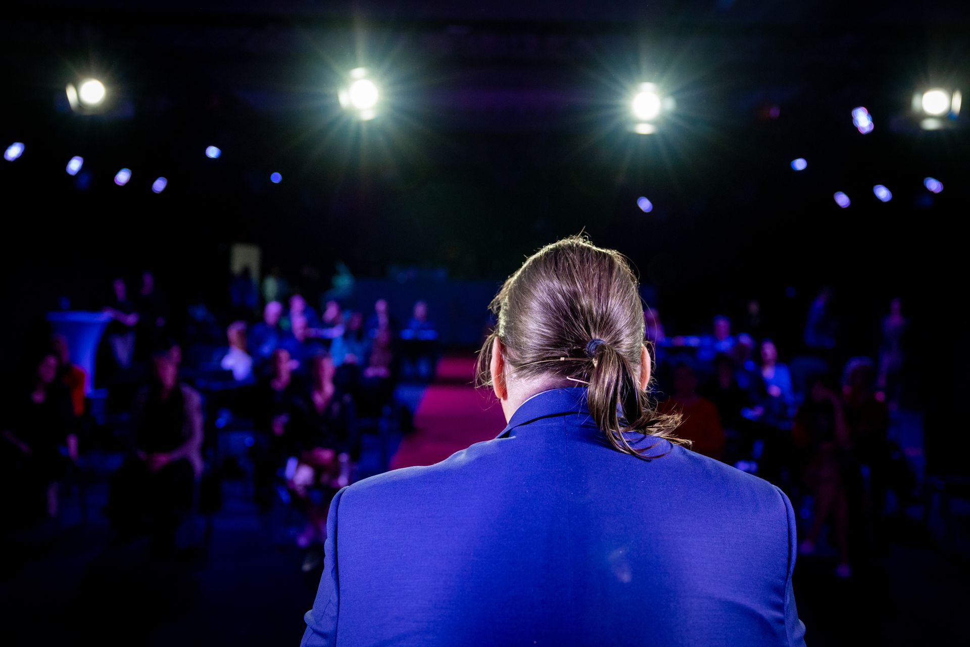 Ein Mann im blauen Blazer steht auf einer hell erleuchteten Bühne und wendet sich dem Publikum zu. Die Zuschauer sitzen in einem dunklen Saal.