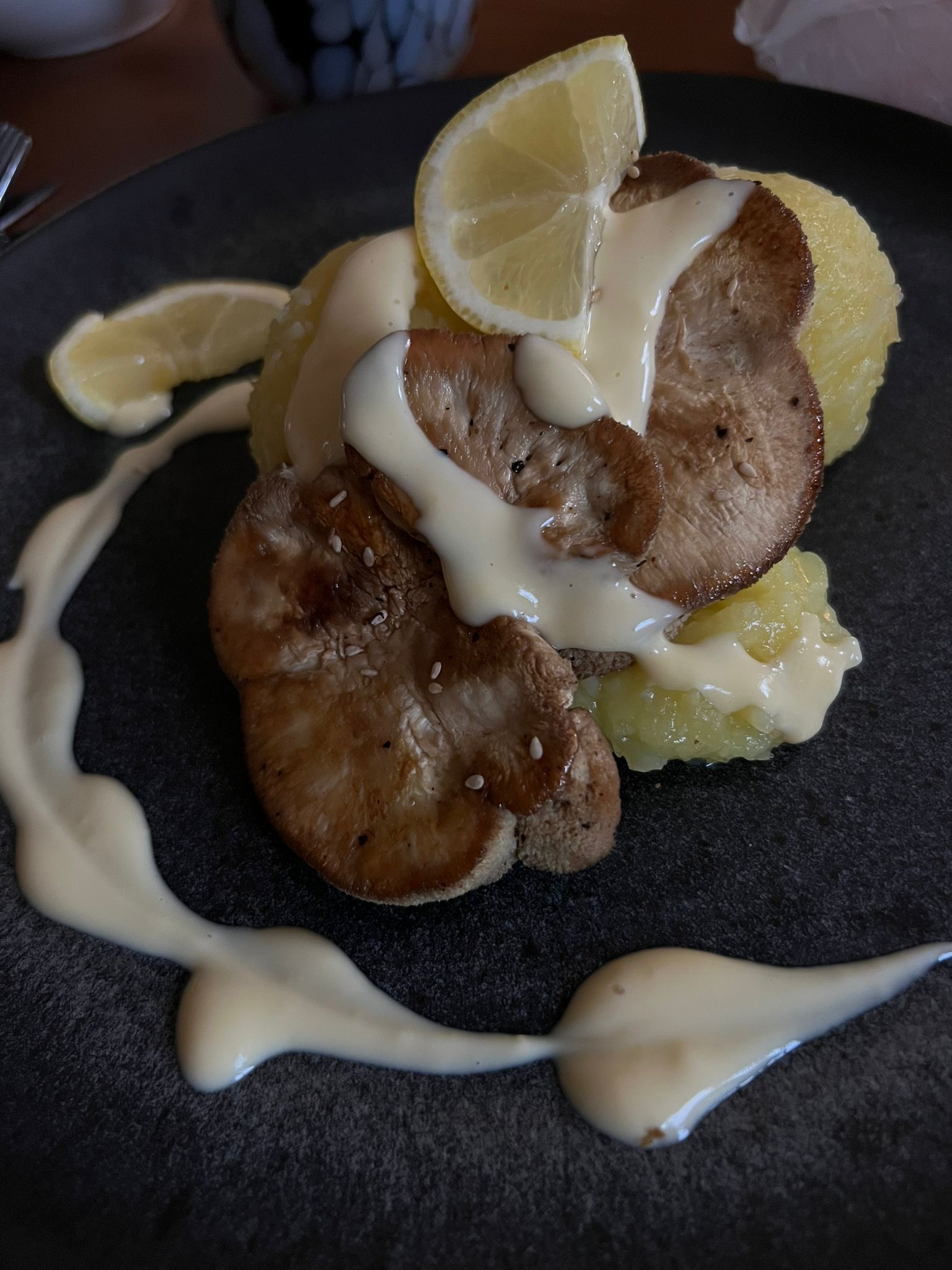 Gebratener Igelstachelbart, Kartoffeln, Zitrone, beträufelt mit weißer Soße, angerichtet auf einem dunklen Teller.