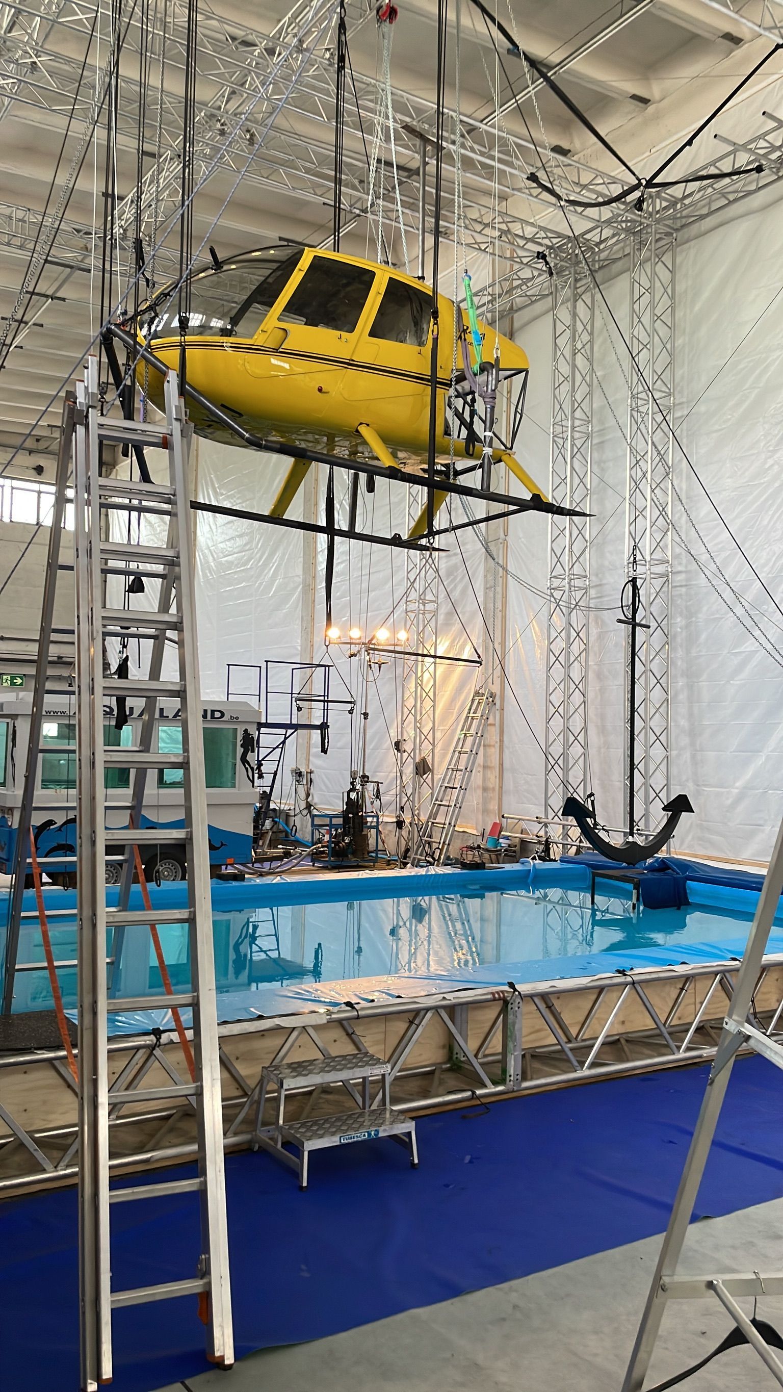 Ein gelber Hubschrauber schwebt über einem Pool in einem Studio, umgeben von Traversen. Im Vordergrund ist eine Leiter zu sehen.