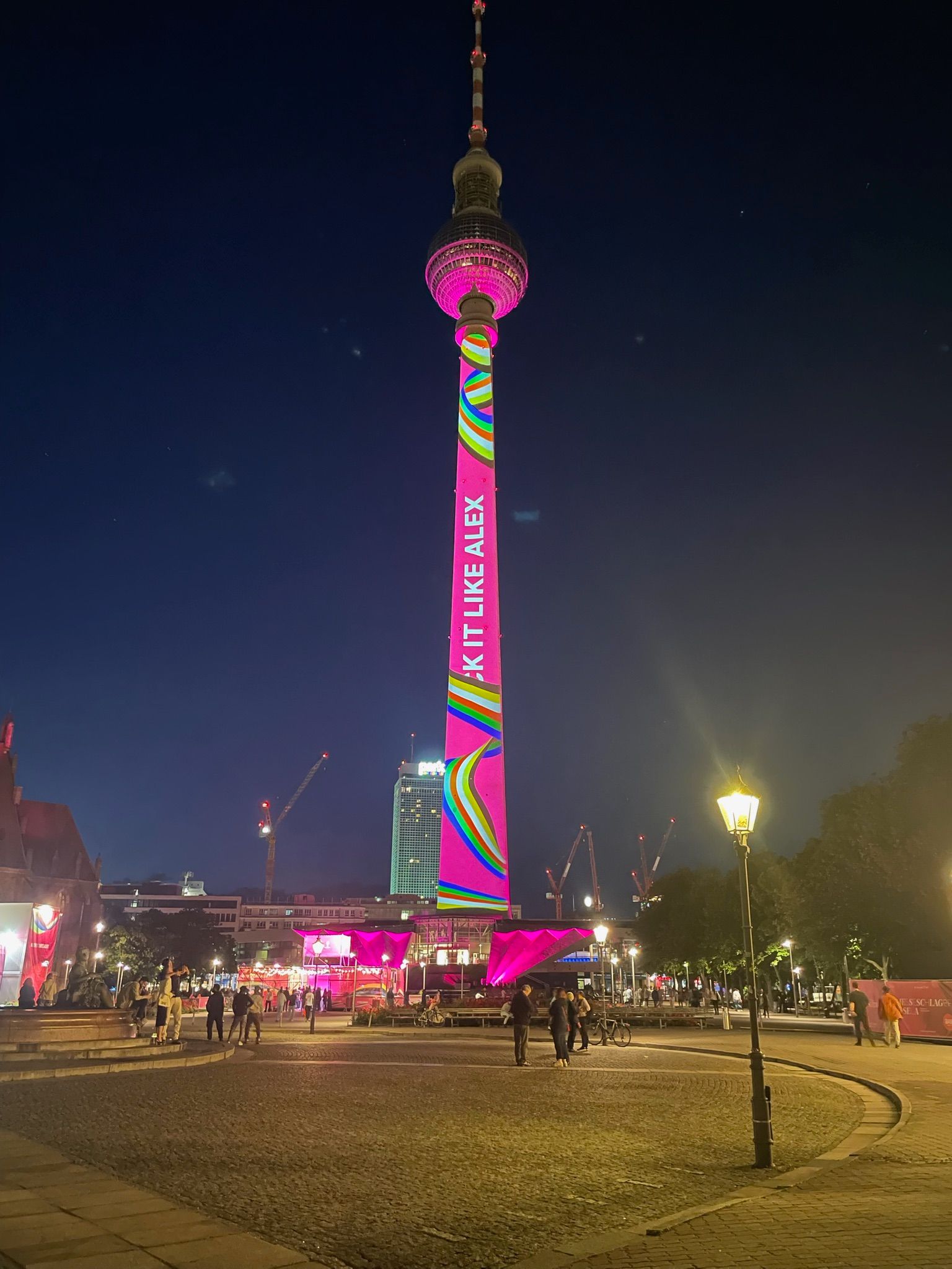 Der Berliner Fernsehturm ist nachts mit bunten Mustern und Schriftzügen beleuchtet. Menschen versammeln sich in der Nähe.