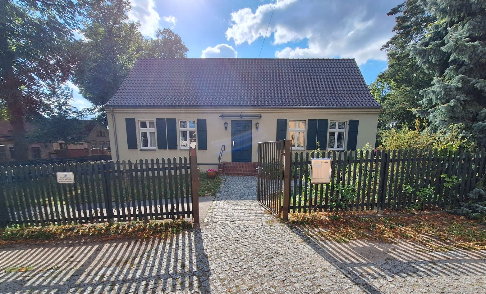 Kleines weißes Häuschen mit blauen Fensterläden hinter einem schwarzen Zaun und einem Kopfsteinpflasterweg, in einem sonnigen Garten.