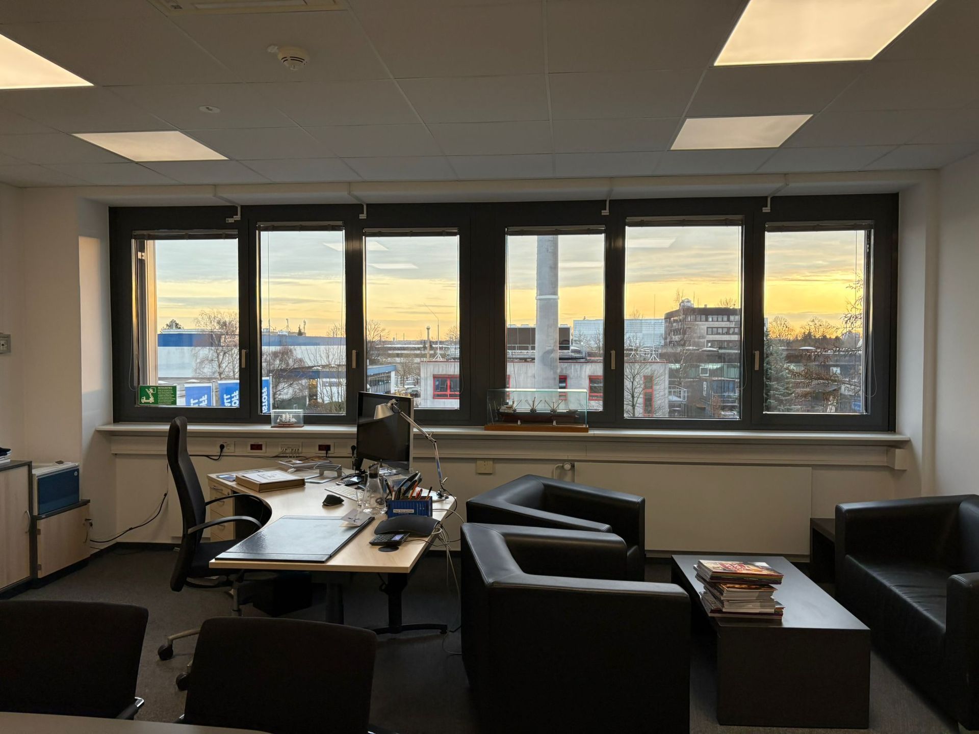 Bürointerieur mit großen Fenstern und Blick auf die Skyline der Stadt bei Sonnenuntergang. Schreibtisch, Stühle und Sofas sind zu sehen.