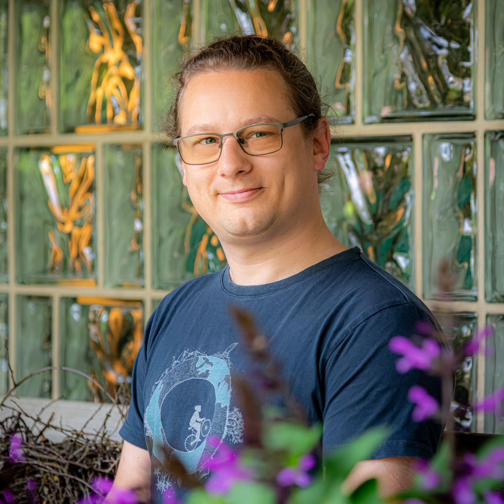 Junger Mann mit Brille und lockigem Haar, lächelnd vor grauem Hintergrund.