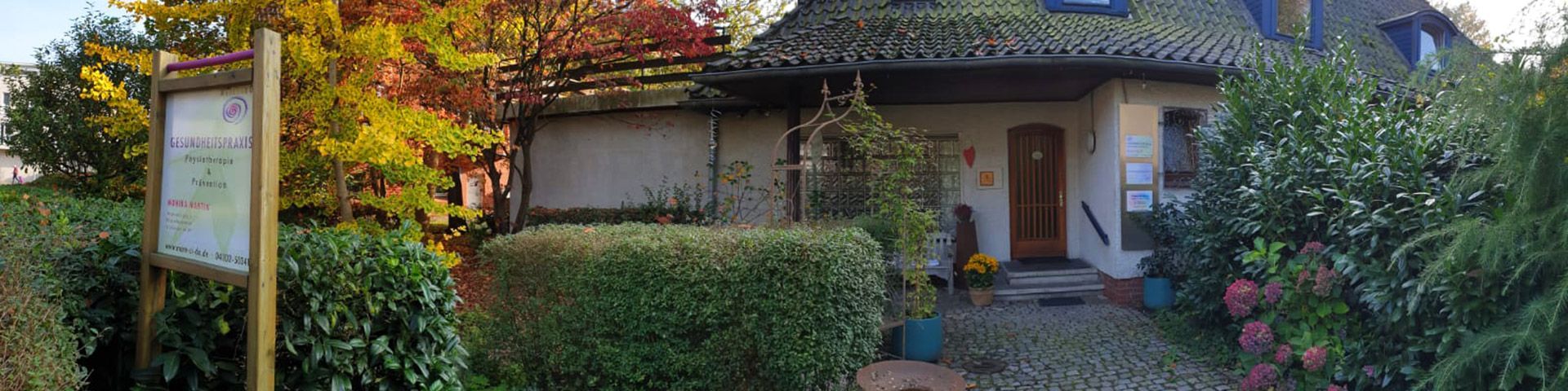 Eine idyllische Gesundheits- und Physiotherapiepraxis mit einem begrünten Eingangsbereich. Ein Holzschild mit Praxisinformationen steht vor herbstlich gefärbtem Laub, während der gepflasterte Weg zum Eingang des Gebäudes führt.