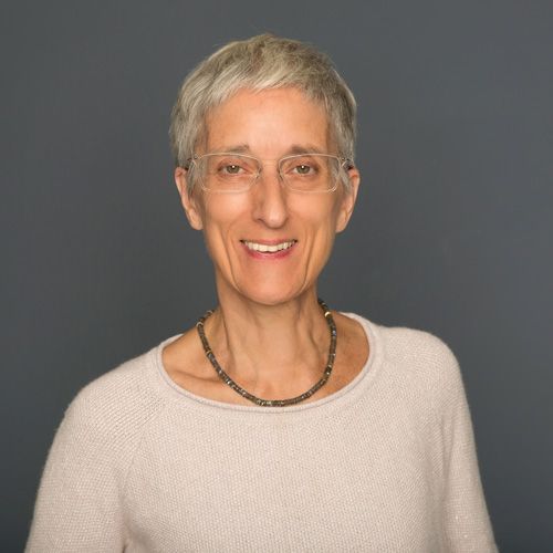 Monika Martin, die Inhaberin der Gesundheits- und Physiotherapiepraxis, lächelt freundlich in die Kamera. Sie trägt eine helle Strickjacke, eine dezente Halskette und eine schlichte Brille. Ihr kurzer, grauer Haarschnitt unterstreicht ihr professionelles und einladendes Auftreten vor einem neutralen, dunklen Hintergrund.