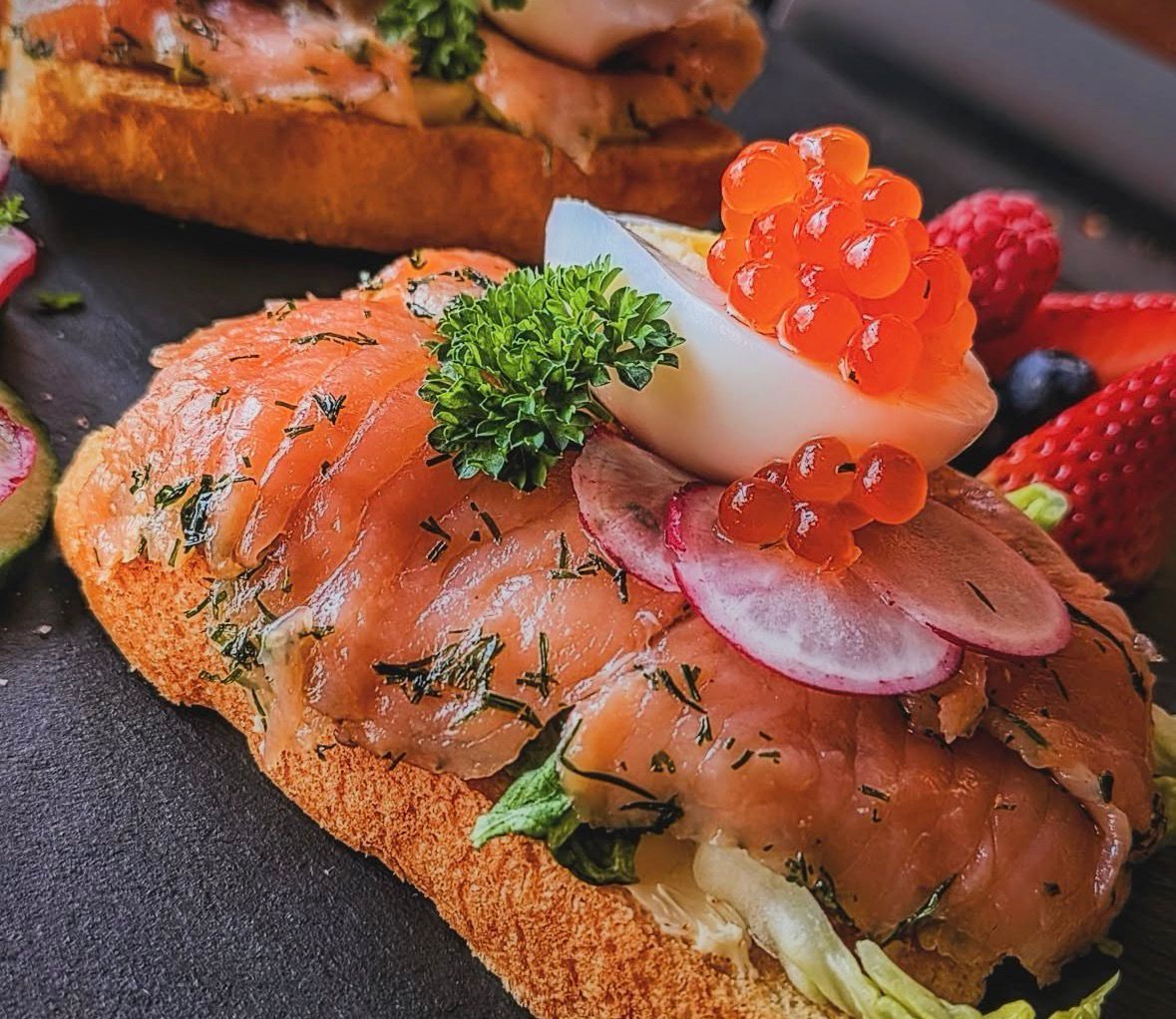 Lachs-Vorspeise auf geröstetem Brot mit Kaviar, Kräutern und Erdbeeren auf einem dunklen Teller