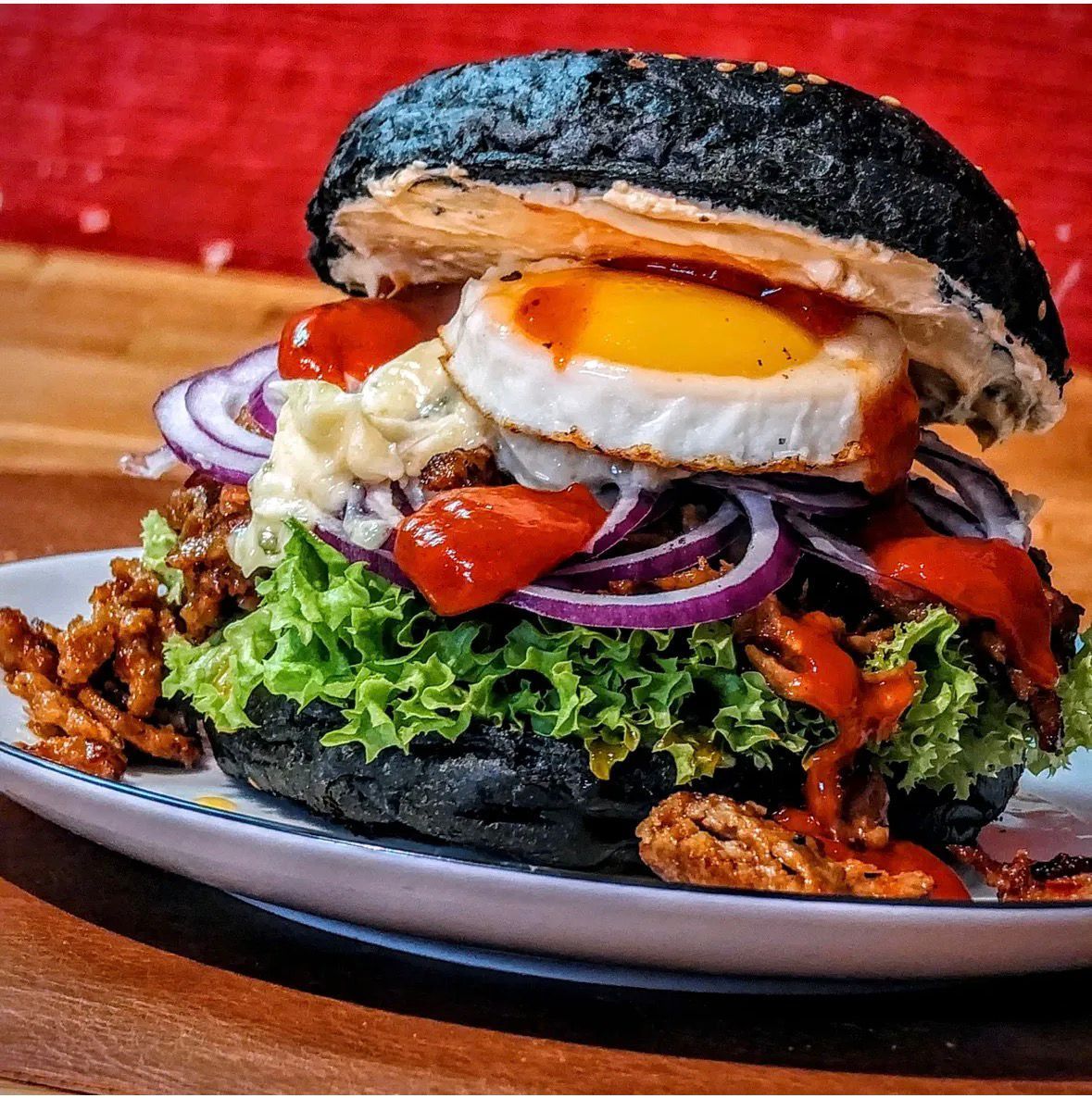 Burger mit schwarzem Brötchen, Spiegelei, Salat, Zwiebeln und Soße auf einem Teller