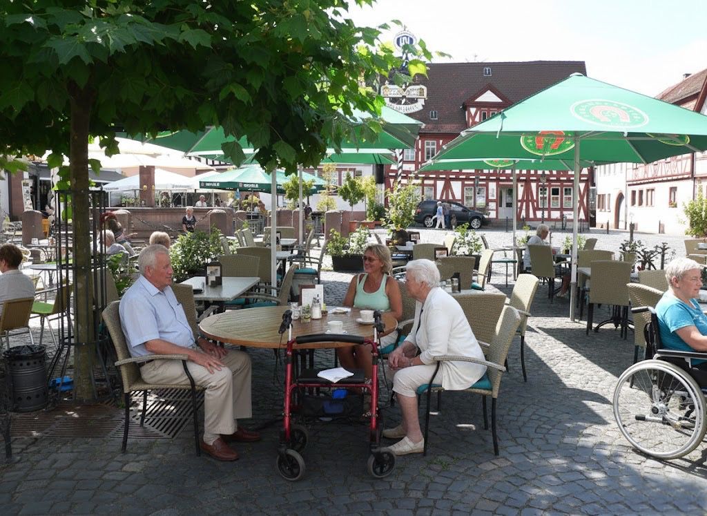 Das Bild zeigt eine entspannte Szene in einem Straßencafé auf dem Marktplatz von Groß-Umstadt. Unter grünen Sonnenschirmen sitzen Senioren und Helfer gemeinsam an einem runden Holztisch. Ein Herr im hellblauen Hemd und zwei Damen sind in ein angeregtes Gespräch vertieft, während vor ihnen Kaffeetassen auf dem Tisch stehen. Im Hintergrund sieht man die malerischen Fachwerkhäuser der Altstadt und weitere Café-Gäste. Auch hier ist die praktische Unterstützung dezent im Bild: Ein roter Rollator steht griffbereit am Tisch. Es ist ein Bild, das zeigt, wie einfach und schön Teilhabe am gesellschaftlichen Leben sein kann, wenn man jemanden an seiner Seite hat.