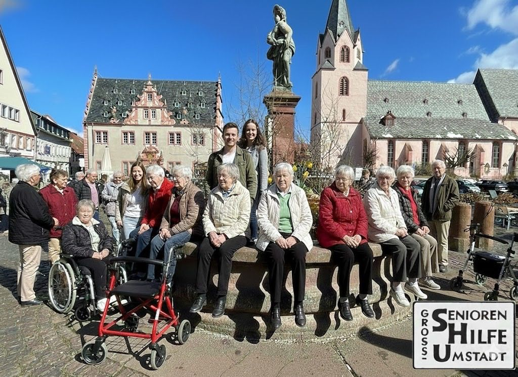 Dieses lebendige Bild zeigt eine Gruppe von Seniorinnen und Senioren, die gemeinsam mit jungen Begleitern am historischen Marktbrunnen von Groß-Umstadt verweilen. Sie sitzen entspannt auf dem Brunnenrand, während im Hintergrund das prächtige Renaissance-Rathaus und die Stadtkirche unter einem strahlend blauen Himmel zu sehen sind. Die Szene wirkt wie ein herzliches Treffen unter Freunden mitten im Stadtleben. Rollstühle und Rollatoren stehen ganz selbstverständlich dabei und zeigen: Dank unserer Unterstützung ist Mobilität kein Hindernis, um die Sonne und das Miteinander auf unserem Marktplatz zu genießen.