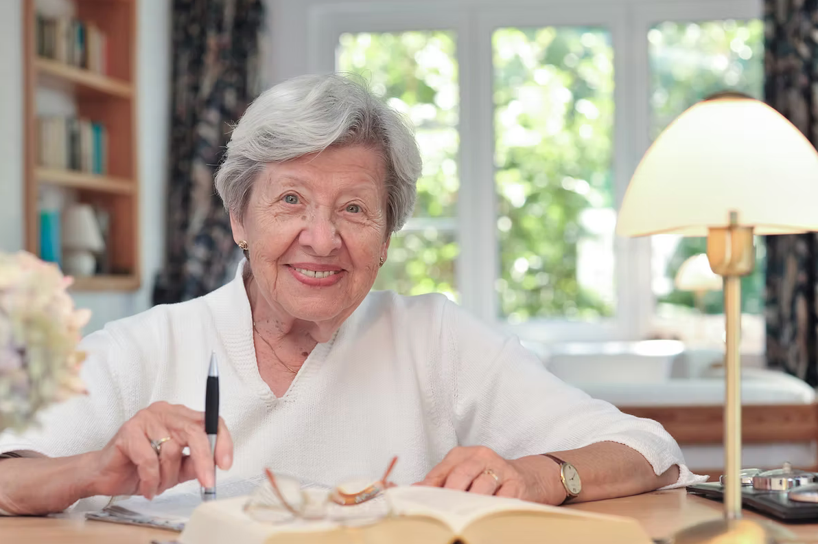 Eine Frau mit grauem Haar lächelt und schreibt an einem Schreibtisch in der Nähe eines Fensters in ein Buch. Lampe und Bücherregal sind zu sehen.